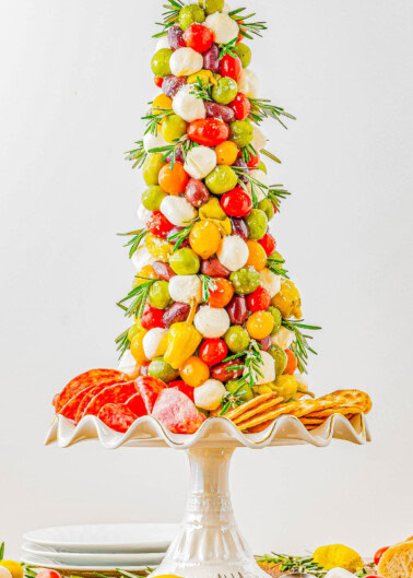 A tree-shaped appetizer display featuring grape tomatoes, mozzarella balls, olives, and peppers on skewers, garnished with rosemary, with salami and crackers at the base on a pedestal stand.