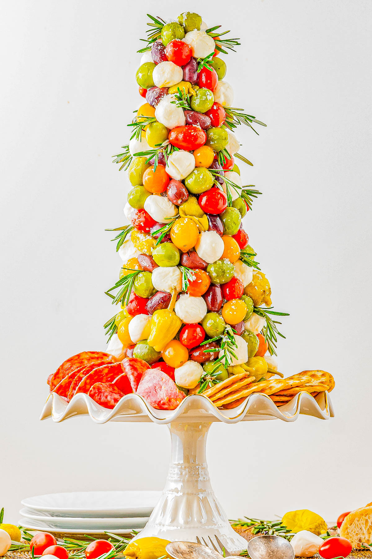 A tree-shaped appetizer display featuring grape tomatoes, mozzarella balls, olives, and peppers on skewers, garnished with rosemary, with salami and crackers at the base on a pedestal stand.