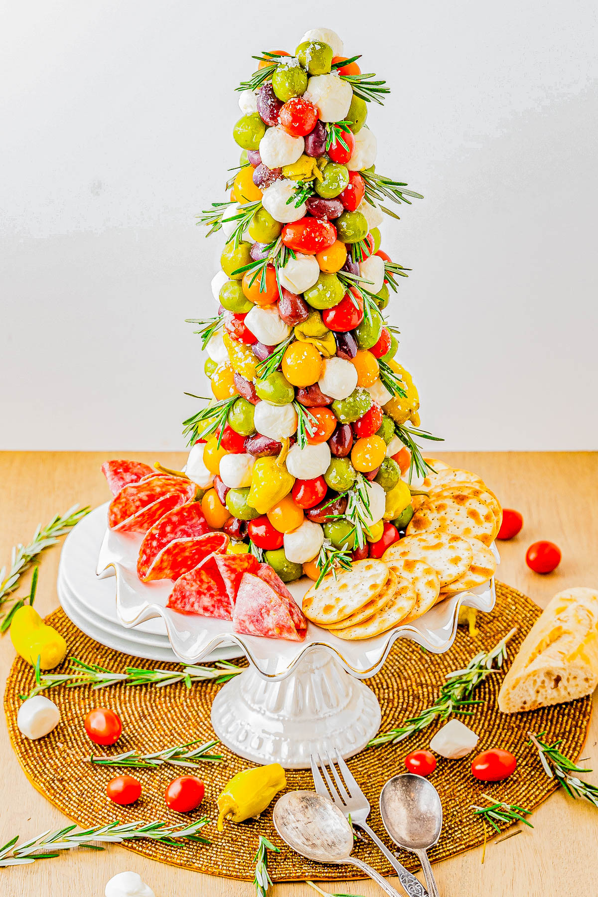 A festive tower of skewered mozzarella, tomatoes, and olives, garnished with rosemary, surrounded by crackers and sliced salami on a white platter.