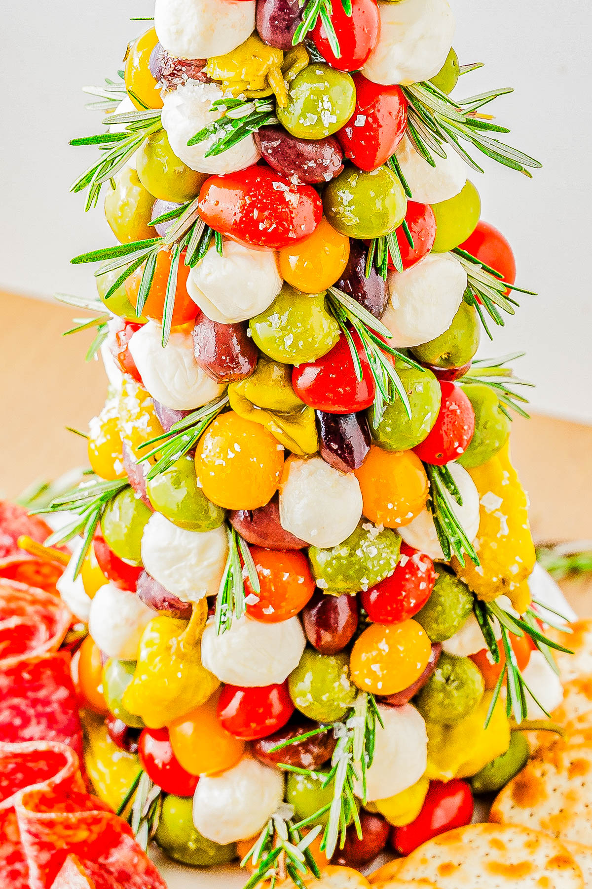 A festive tower of colorful cherry tomatoes, cheese balls, and olives, garnished with rosemary, next to slices of cured meat and crackers.