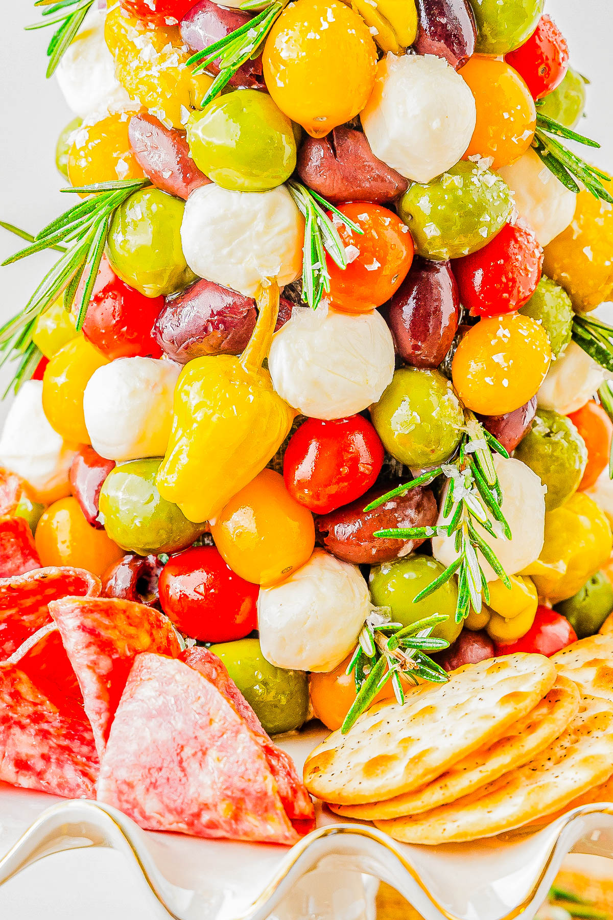 A decorative arrangement of mixed olives, mozzarella balls, and peppers stacked in a cone shape, garnished with rosemary, surrounded by slices of salami and crackers on a platter.