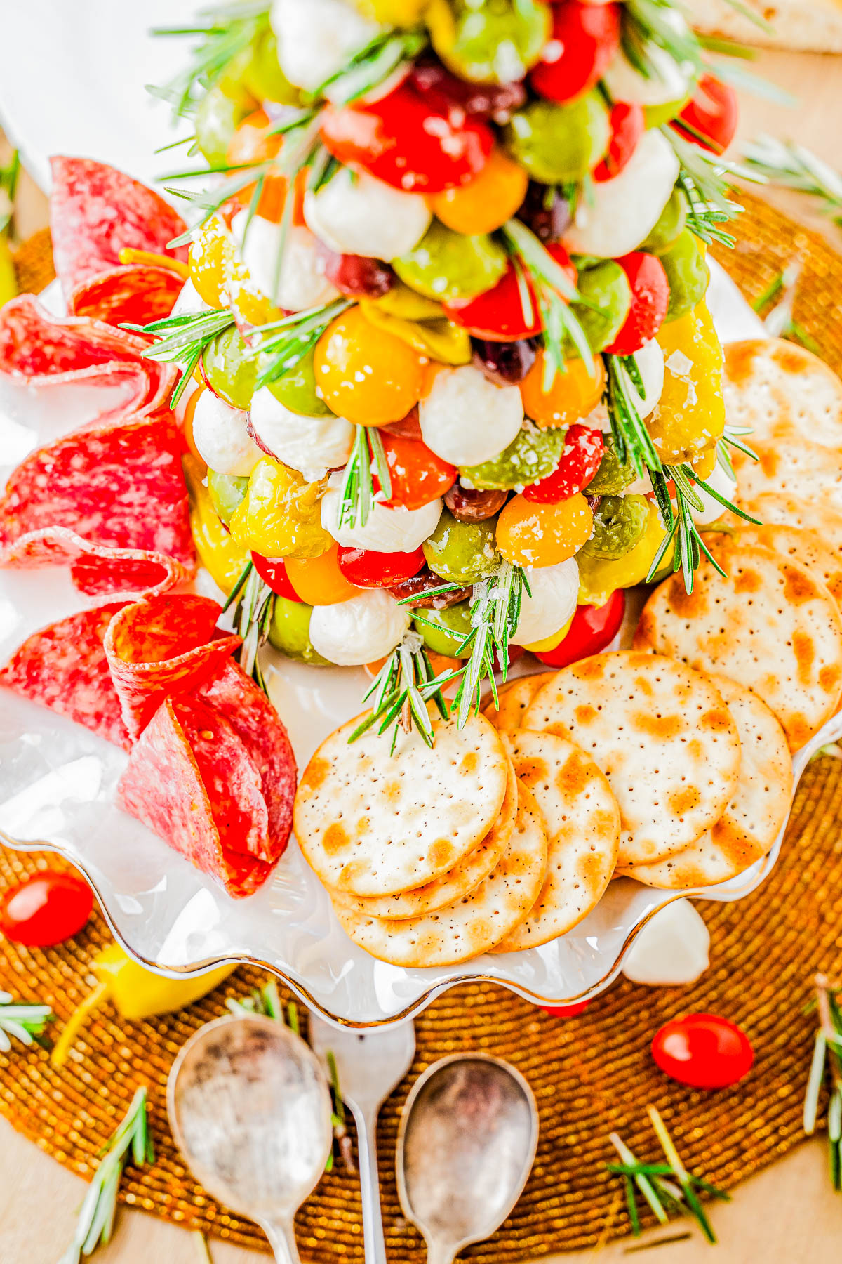 A charcuterie display with a tree-shaped arrangement of cheese and tomatoes, surrounded by crackers, salami, and garnished with rosemary, set on a white platter.