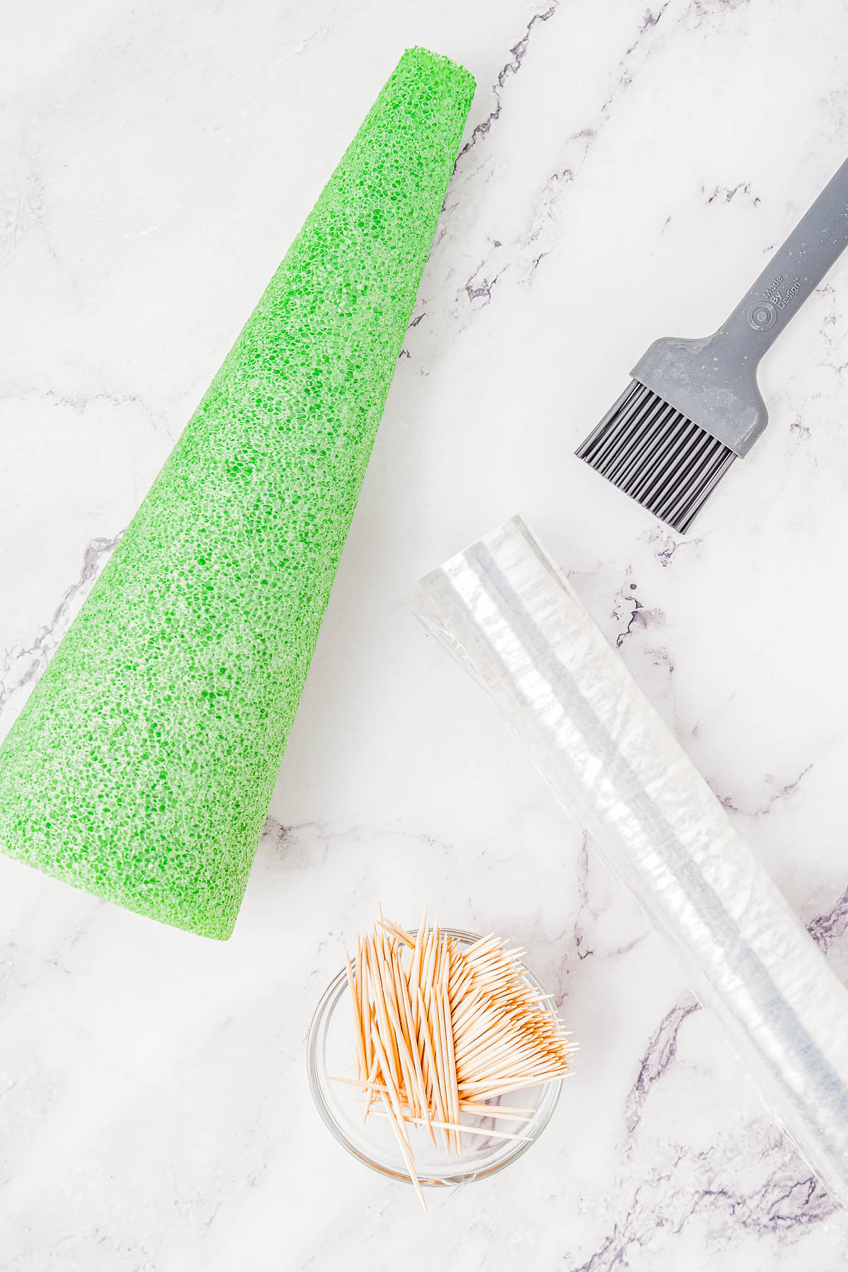 Green foam cone, gray meat tenderizer, bowl of toothpicks, and roll of plastic wrap on marble surface.