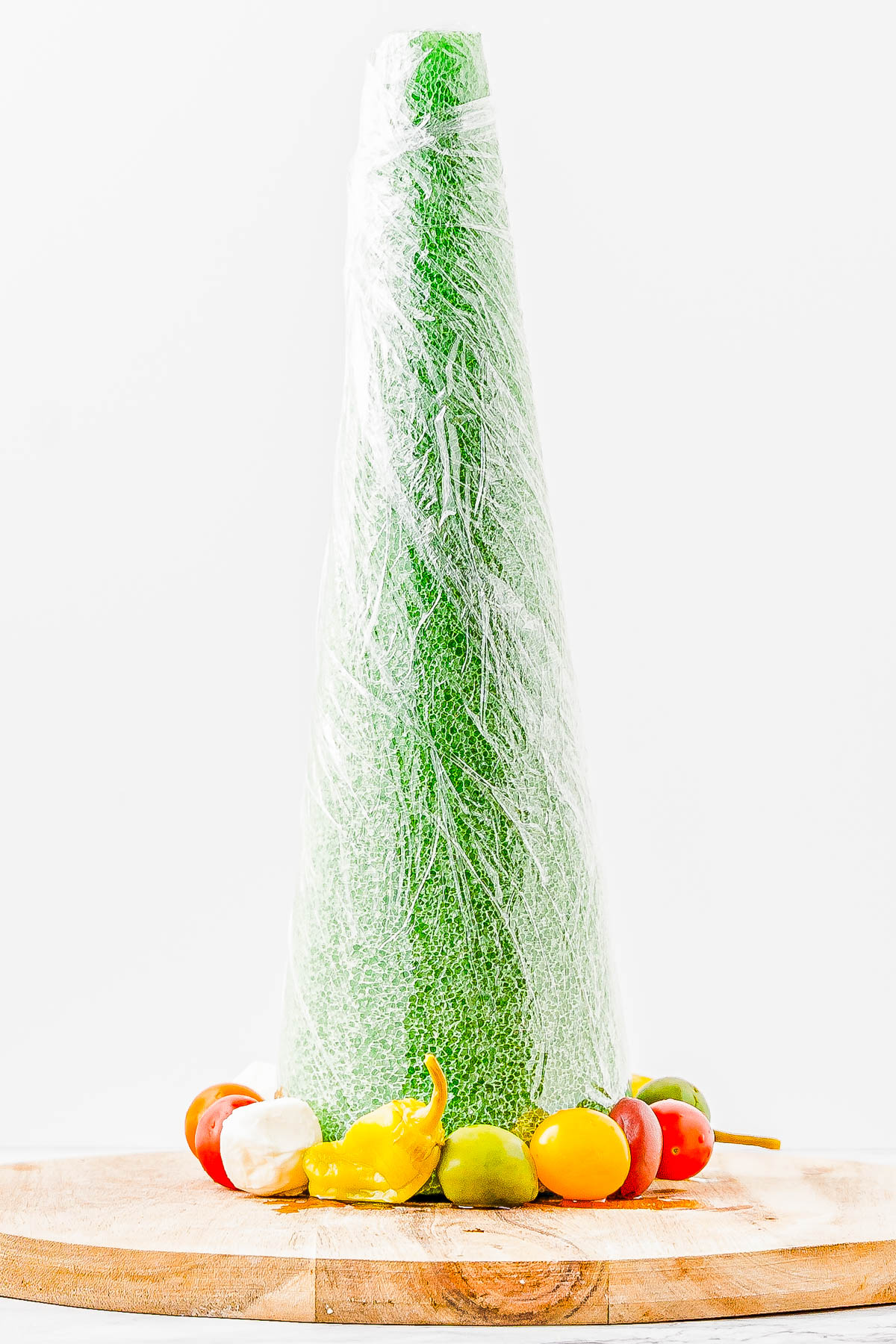 A green foam cone wrapped in plastic sits on a wooden surface, surrounded by various small colorful peppers.