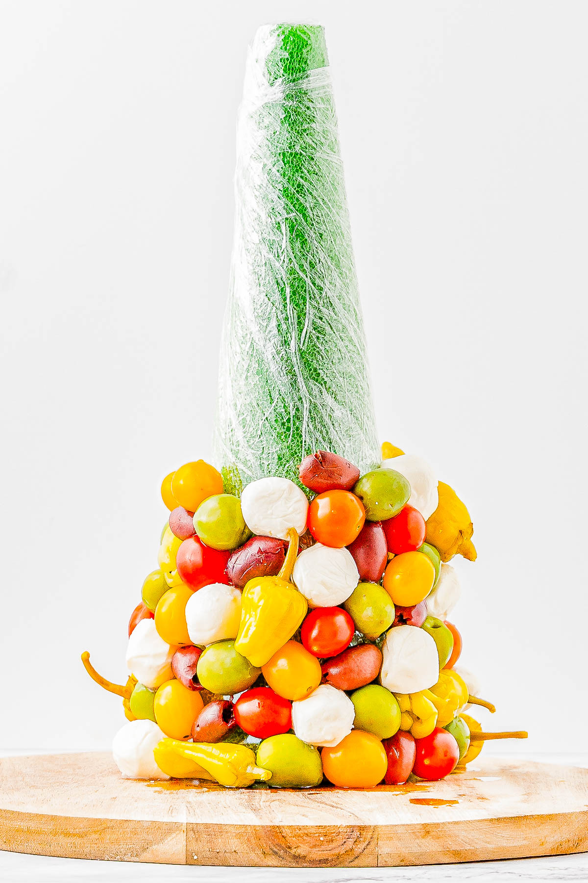 A tall cone wrapped in plastic is decorated with colorful cherry tomatoes, mozzarella balls, and yellow peppers, placed on a wooden board.