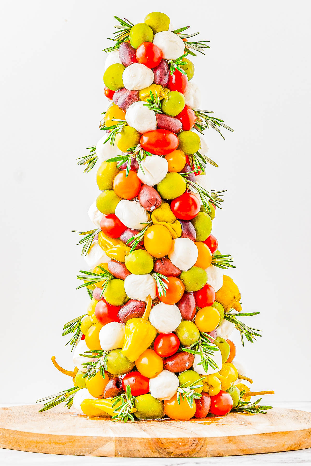 A colorful, cone-shaped tower of cherry tomatoes, mozzarella balls, olives, and peppers, garnished with rosemary sprigs on a round wooden board.