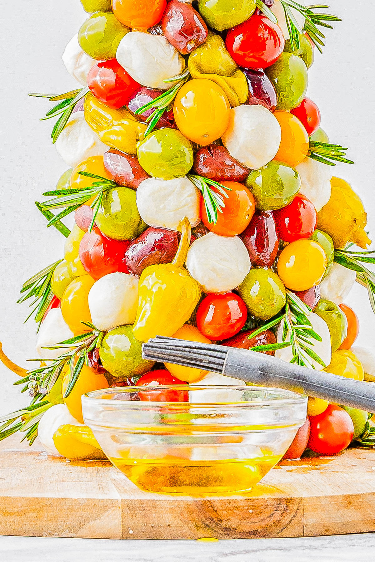 A vertical skewer with assorted colored olives, mozzarella balls, and cherry tomatoes garnished with rosemary. A small bowl of olive oil with a brush is in the foreground.