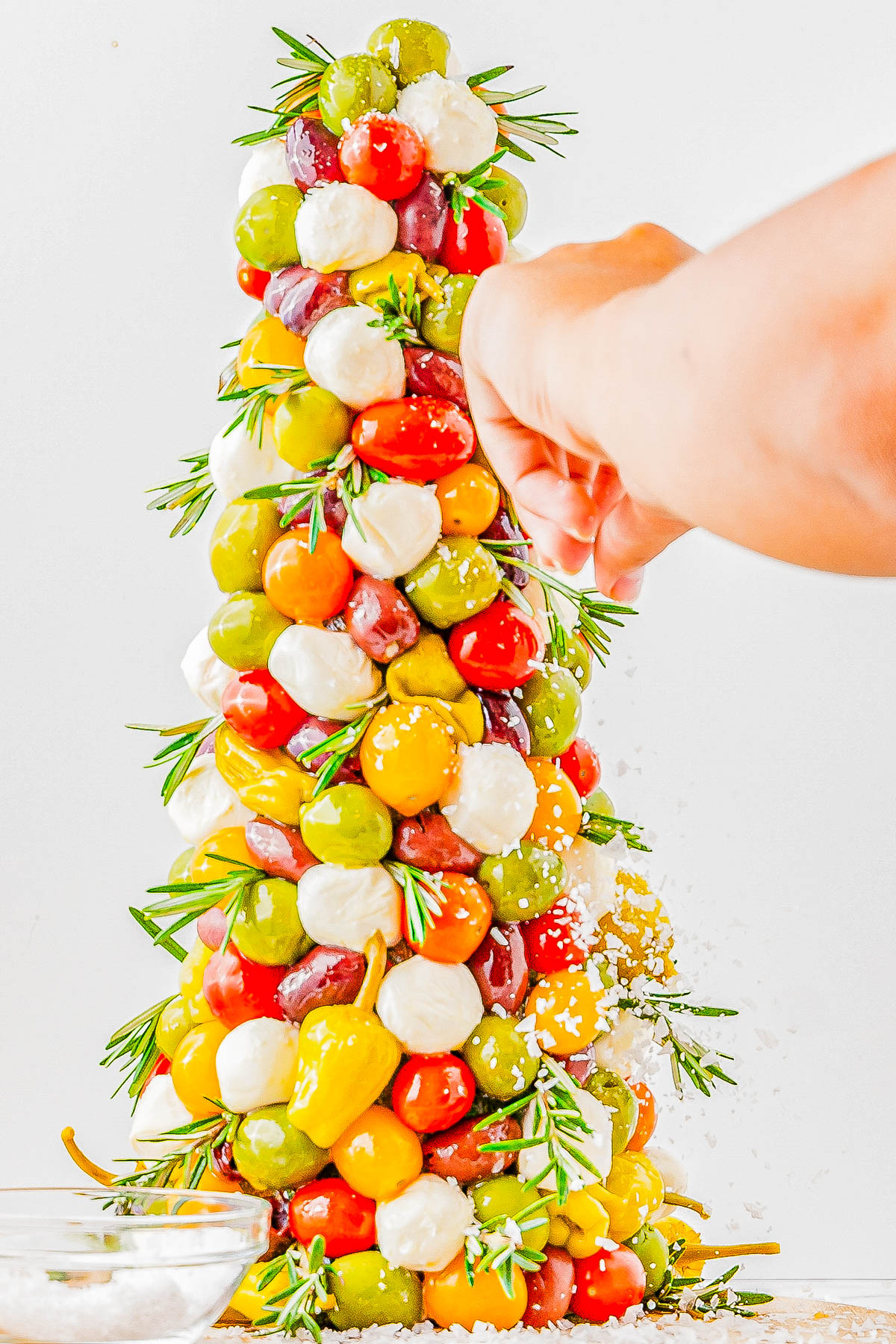 A hand sprinkles cheese onto a tower made of colorful olives, cherry tomatoes, and mozzarella balls, garnished with rosemary sprigs.