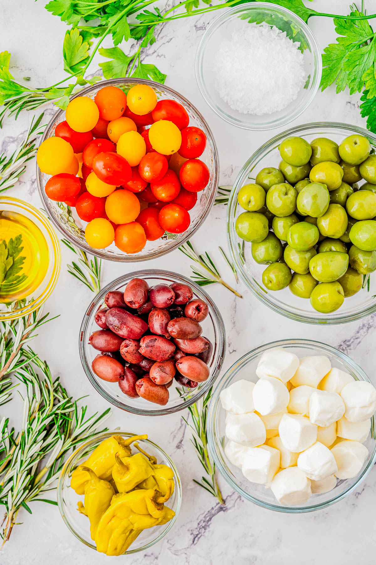 Bowls of cherry tomatoes, green olives, kalamata olives, mozzarella cheese, pepperoncini, salt, and olive oil arranged with rosemary and parsley on a marble surface.