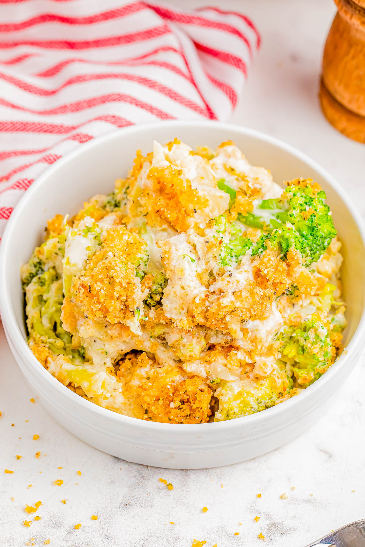 A bowl of broccoli and cauliflower casserole topped with golden breadcrumbs, placed on a marble surface with a striped cloth nearby.