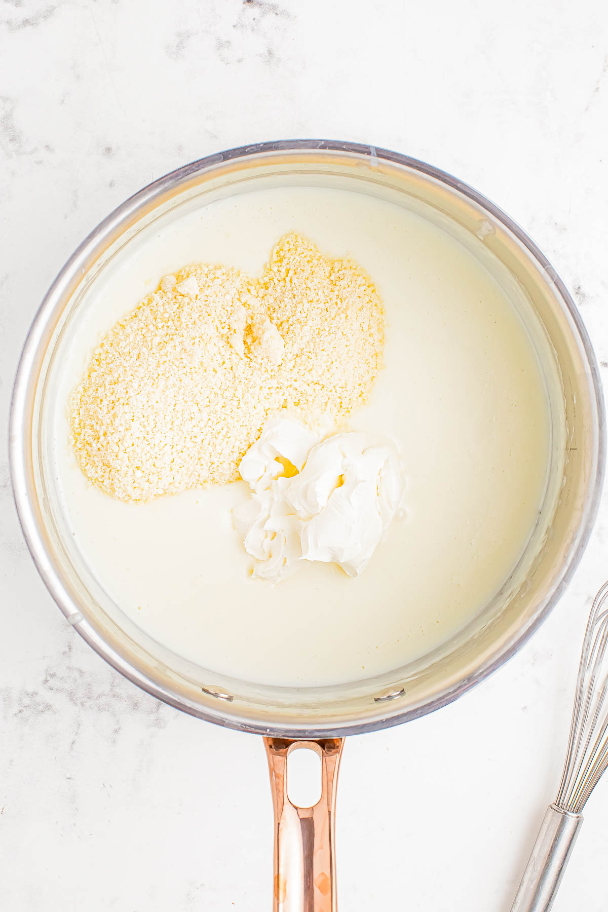 A saucepan with creamy liquid, cornmeal, and a dollop of white cream. A whisk is beside the pan.