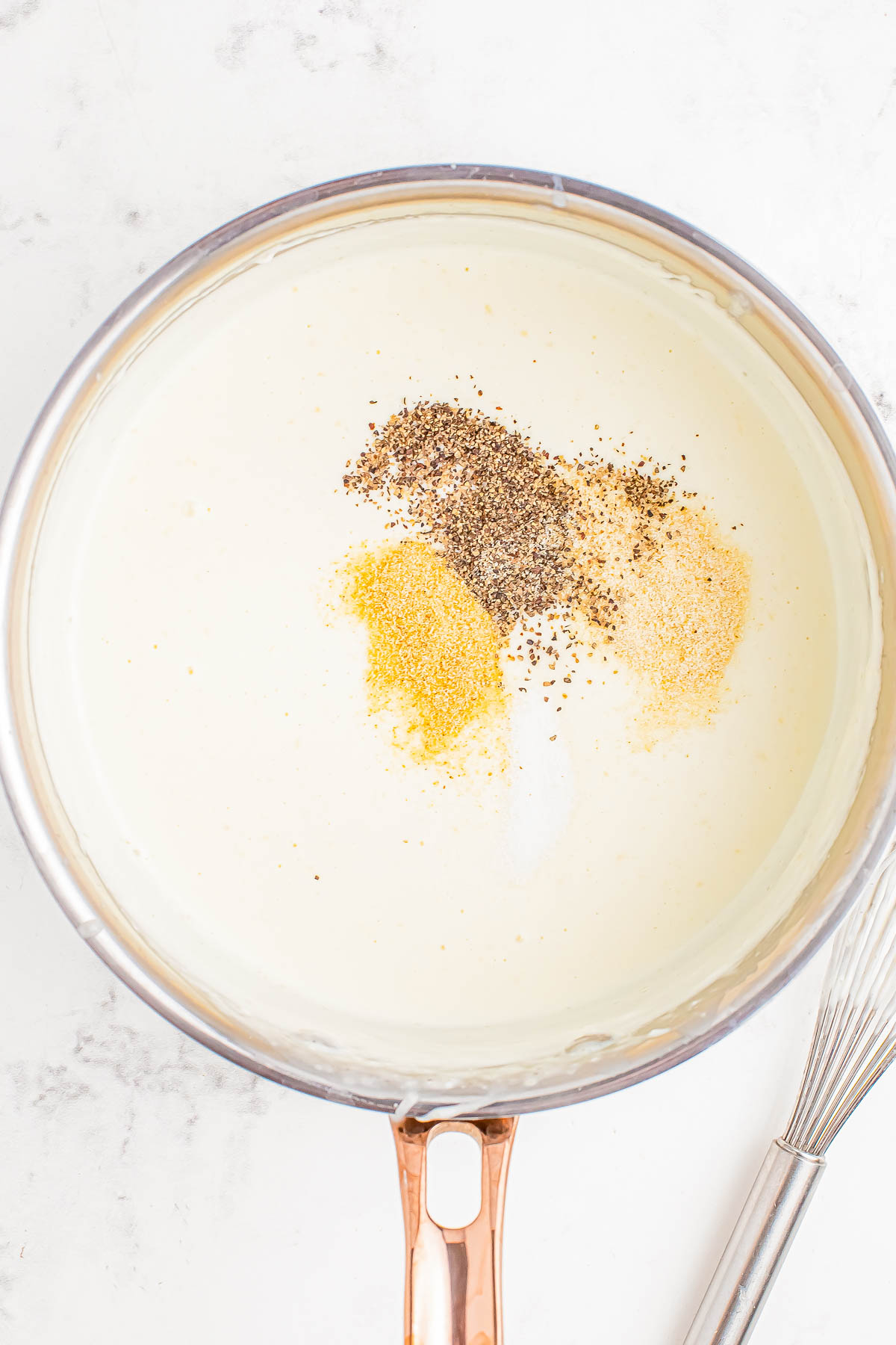 A saucepan of creamy white sauce with black pepper, garlic powder, and onion powder on top.