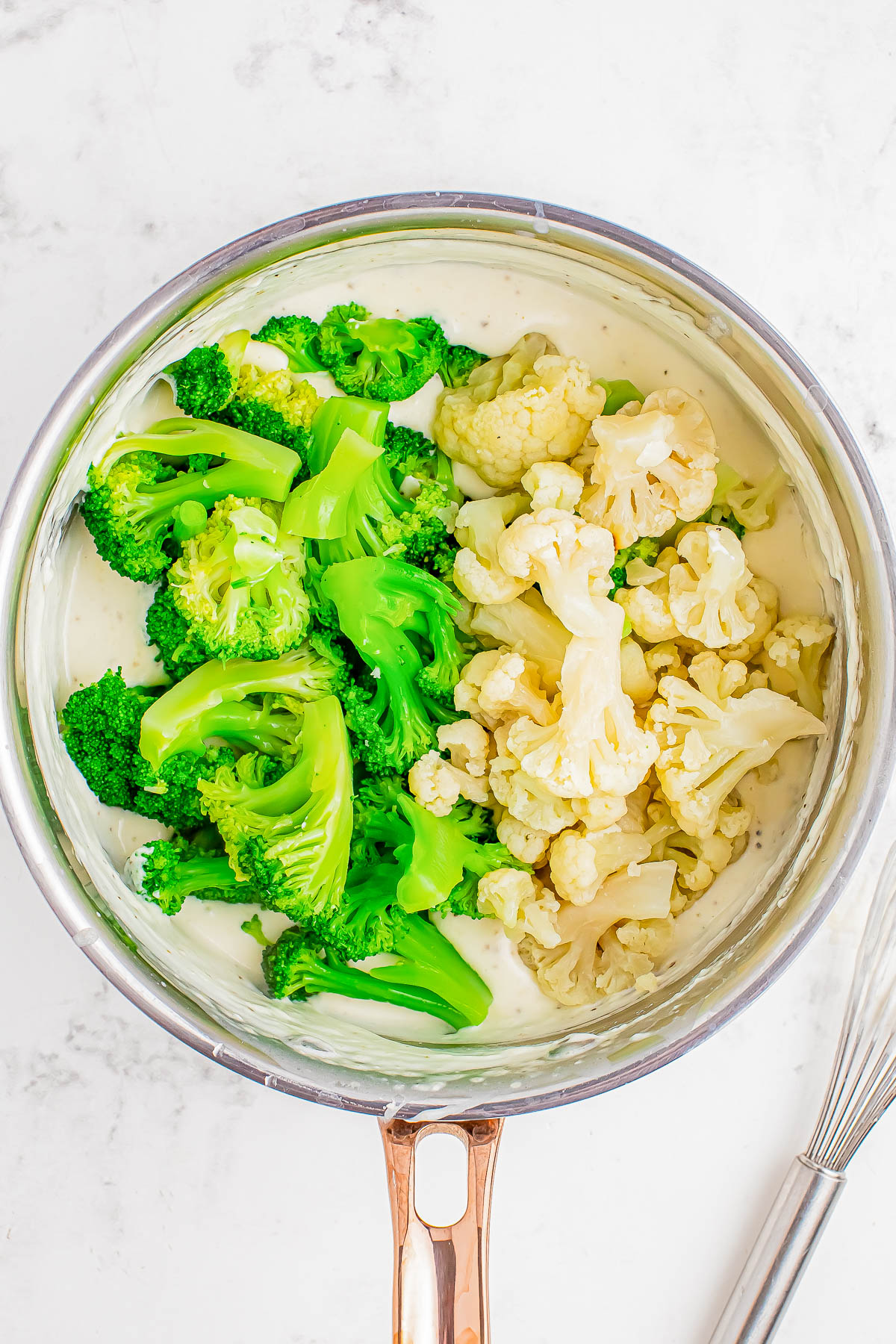 A saucepan with steamed broccoli and cauliflower on a creamy white sauce, placed on a marble surface.