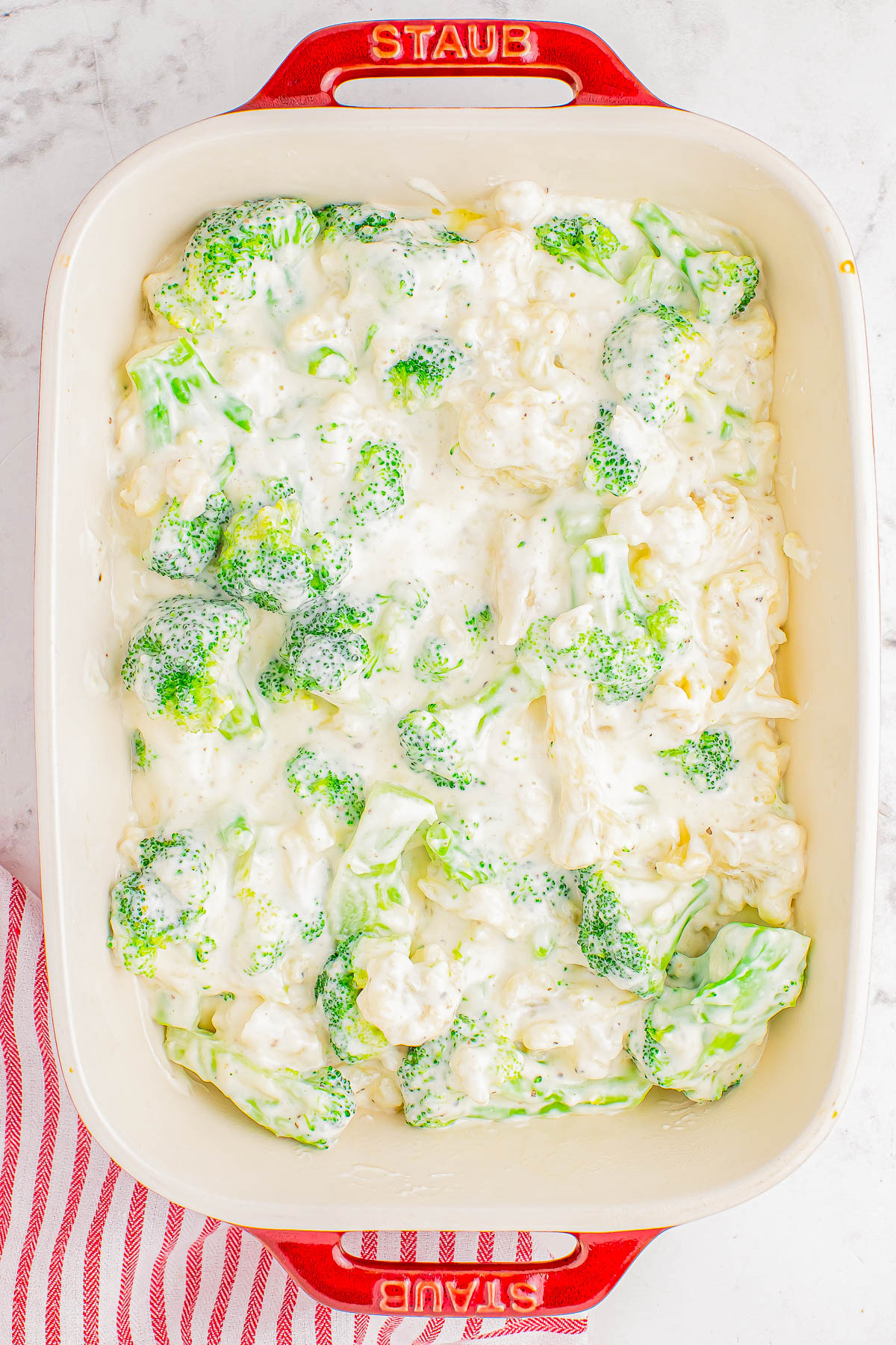 A red-bordered baking dish filled with a mixture of broccoli florets and creamy white sauce, set on a striped red and white cloth.