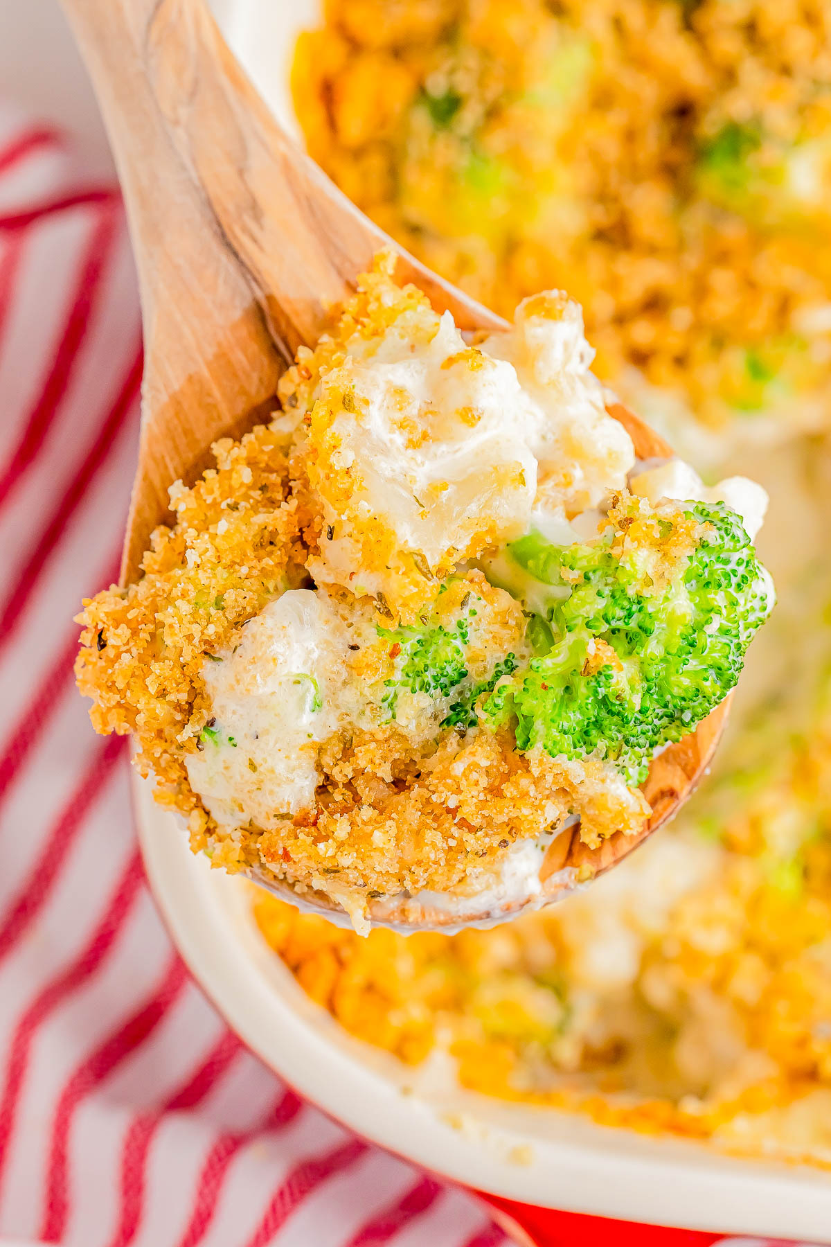 A wooden spoon holds a portion of broccoli and cheese casserole with a crispy breadcrumb topping, above a red and white striped cloth.