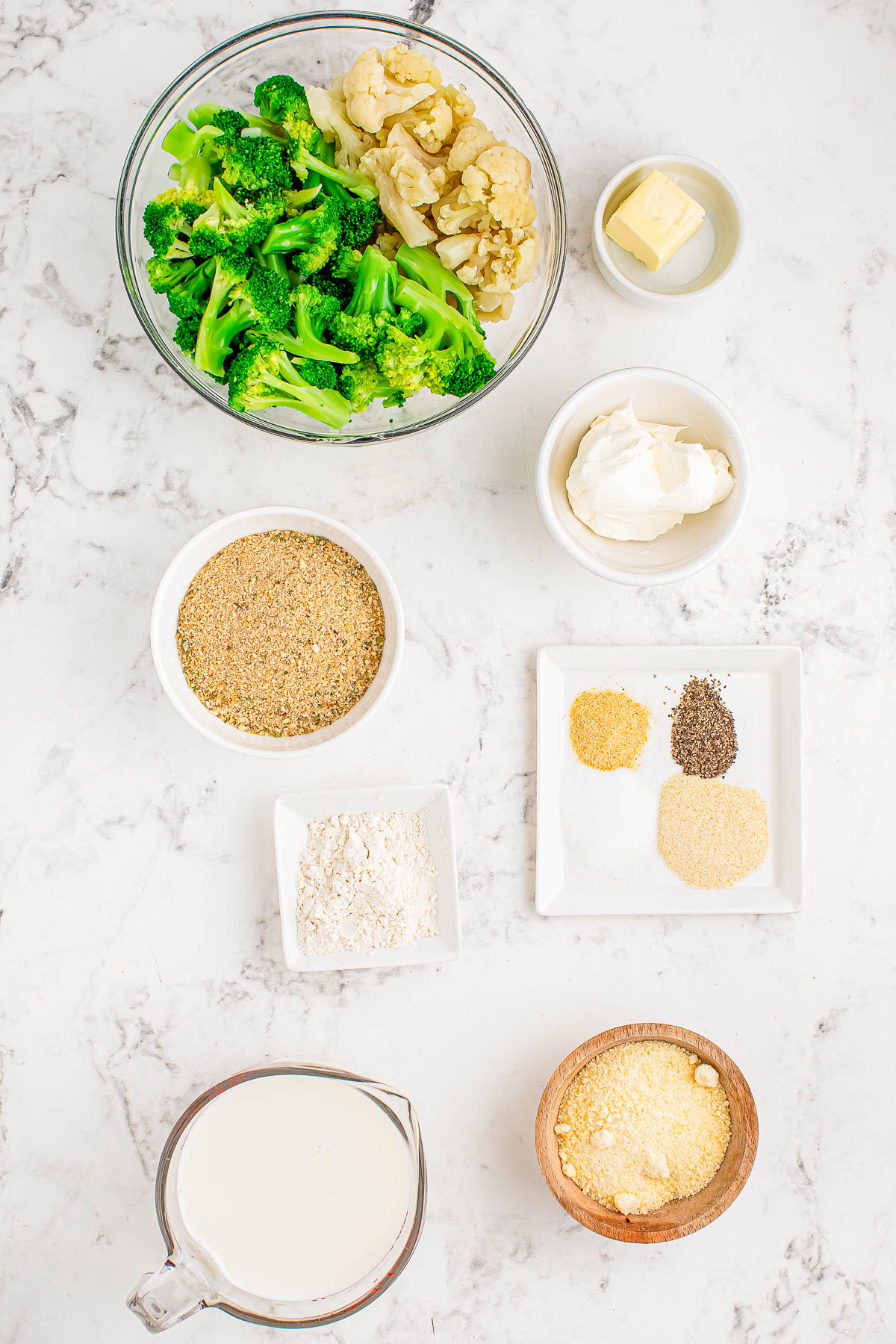 Ingredients on a marble countertop: broccoli, cauliflower, butter, cream, breadcrumbs, flour, spices, milk, and cornmeal in separate bowls.