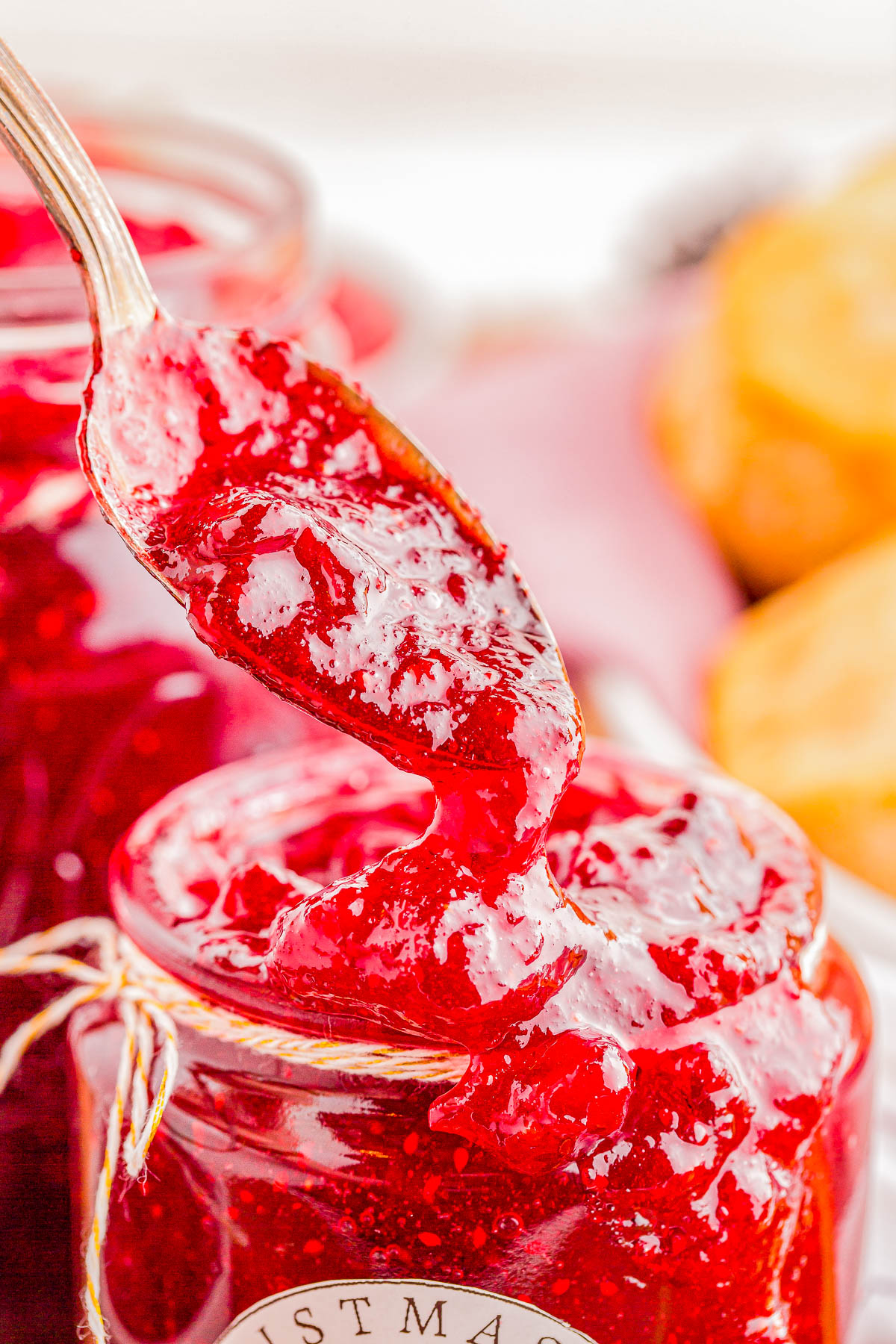 Close-up of a spoon scooping bright red jam from a glass jar. The jar is wrapped with twine and has a blurred background.