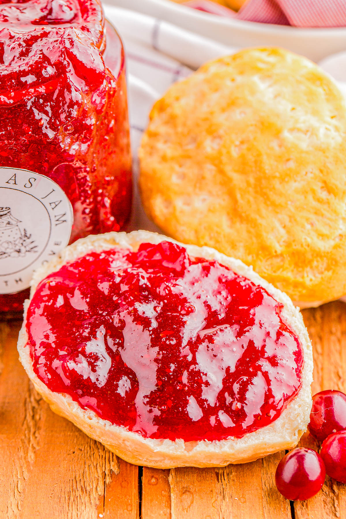 A jar of red jam next to a biscuit topped with jam, a plain biscuit, and a few cranberries on a wooden surface.