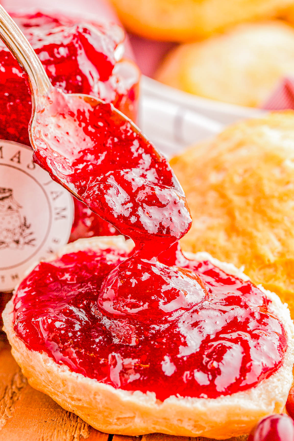 A spoon spreads bright red strawberry jam on a halved biscuit. A jar of jam and more biscuits are blurred in the background.