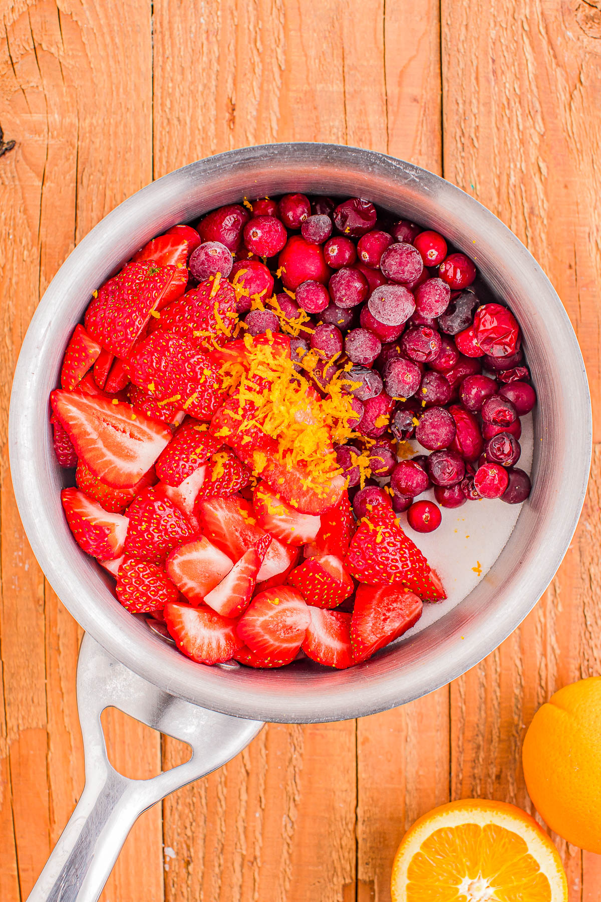 A saucepan filled with sliced strawberries, whole cranberries, and orange zest on a wooden surface. An orange is partially visible beside the saucepan.