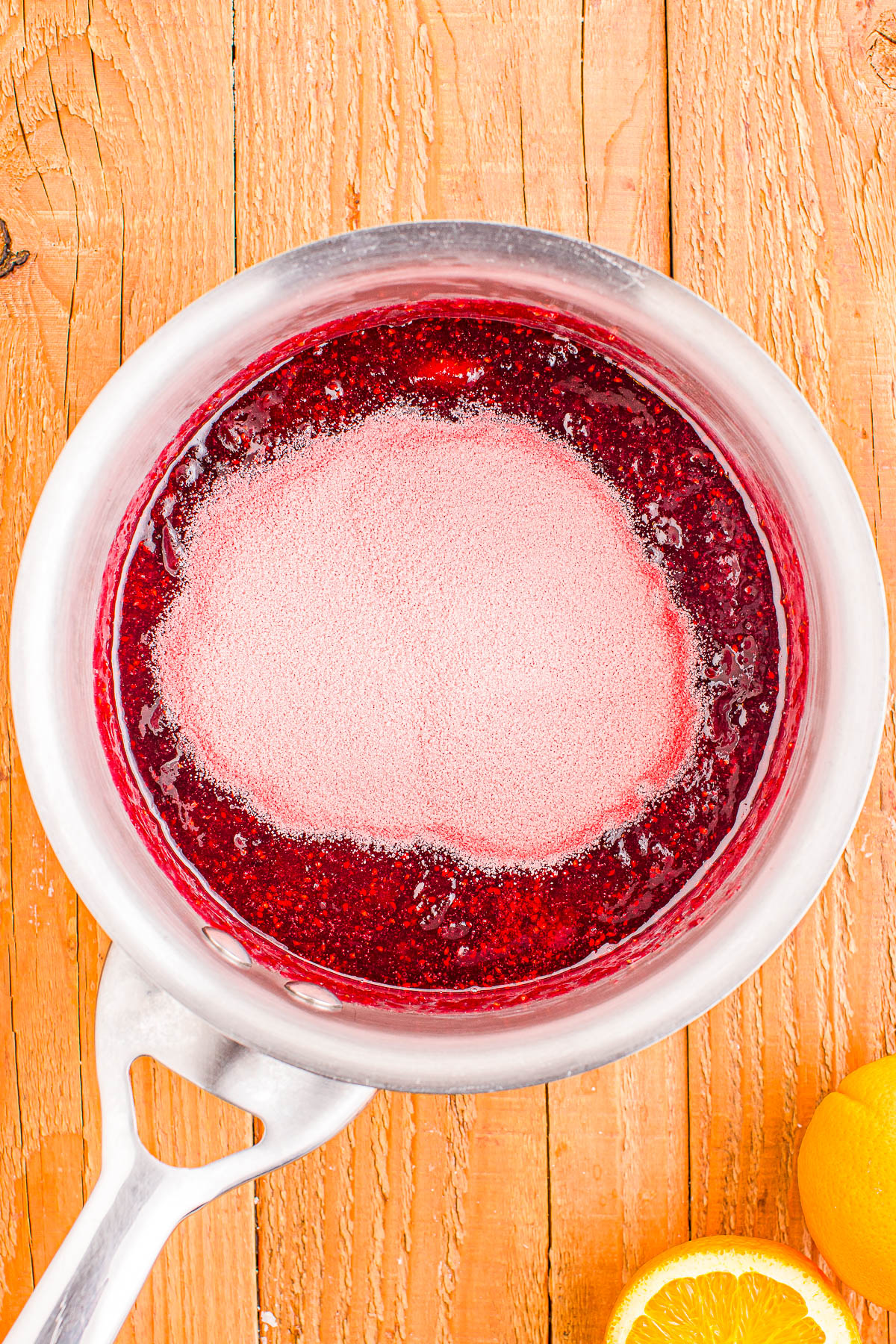 A saucepan filled with red liquid and a layer of white powder on top, placed on a wooden surface with a sliced lemon nearby.