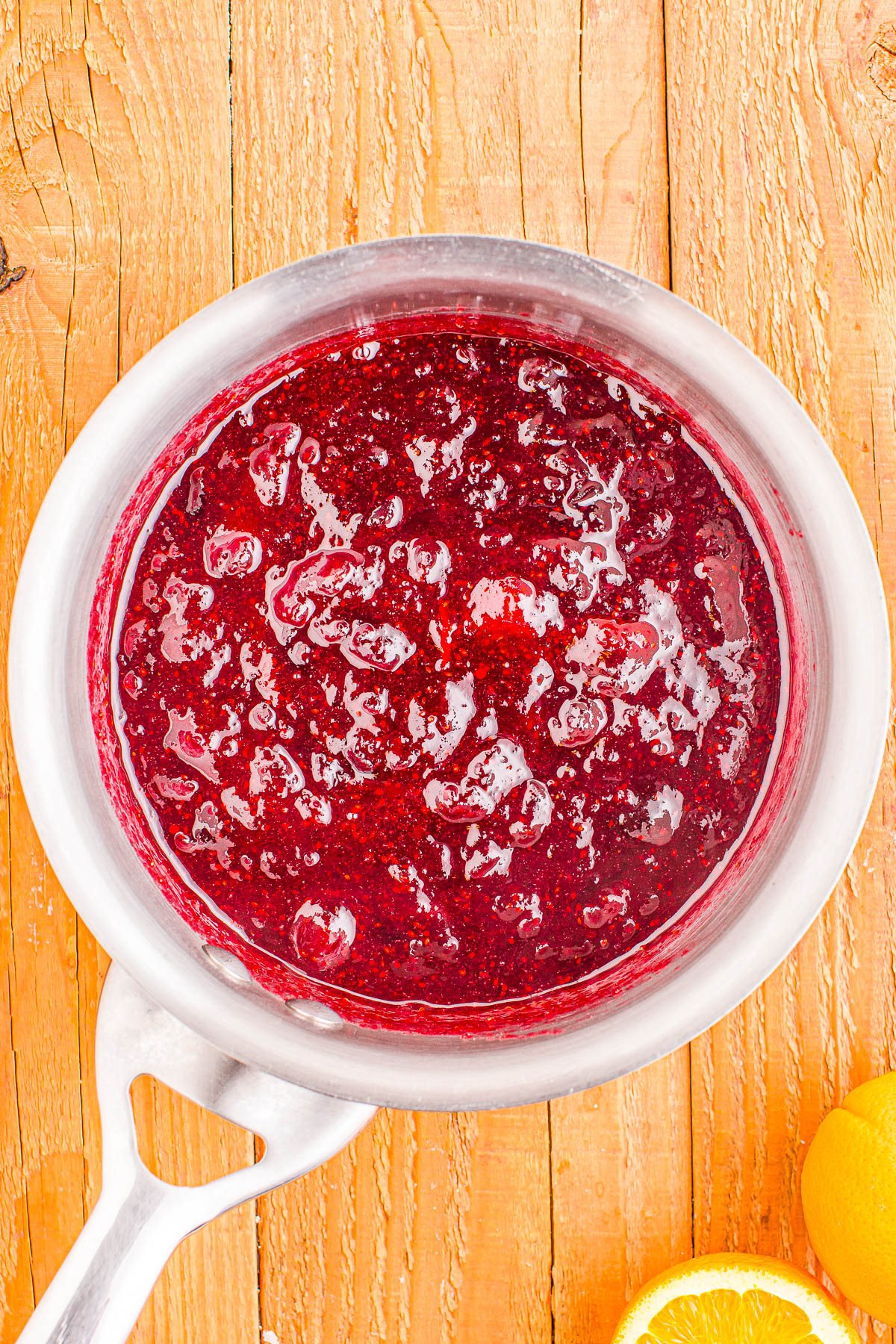 A saucepan containing a bubbling mixture of red berry sauce rests on a wooden surface. An orange slice is nearby.