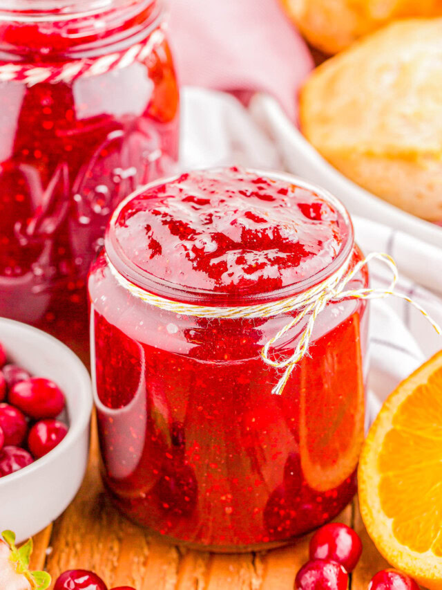 Jar of red jam next to fresh cranberries and an orange slice on a wooden surface, with a larger jar of jam and pastries in the background.