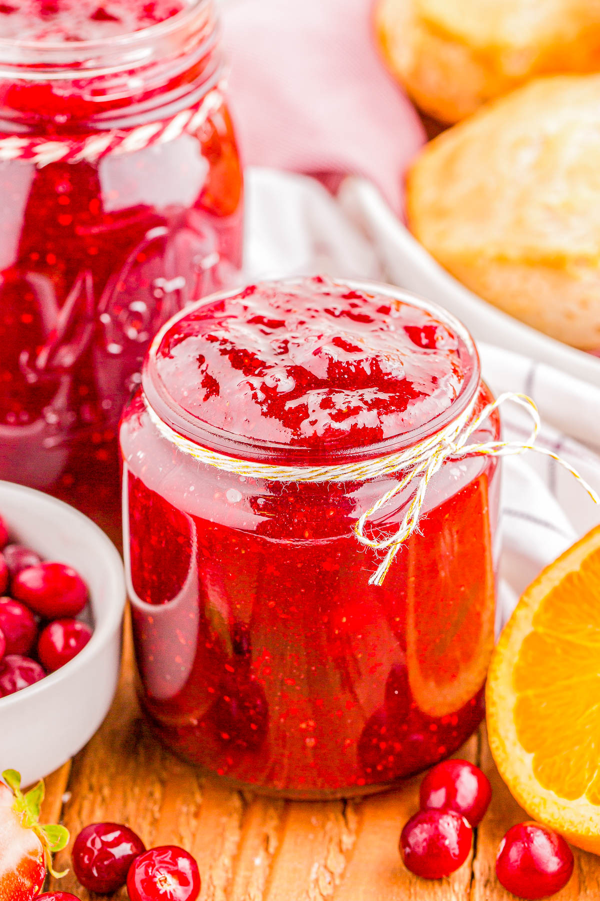 Jar of red jam next to fresh cranberries and an orange slice on a wooden surface, with a larger jar of jam and pastries in the background.