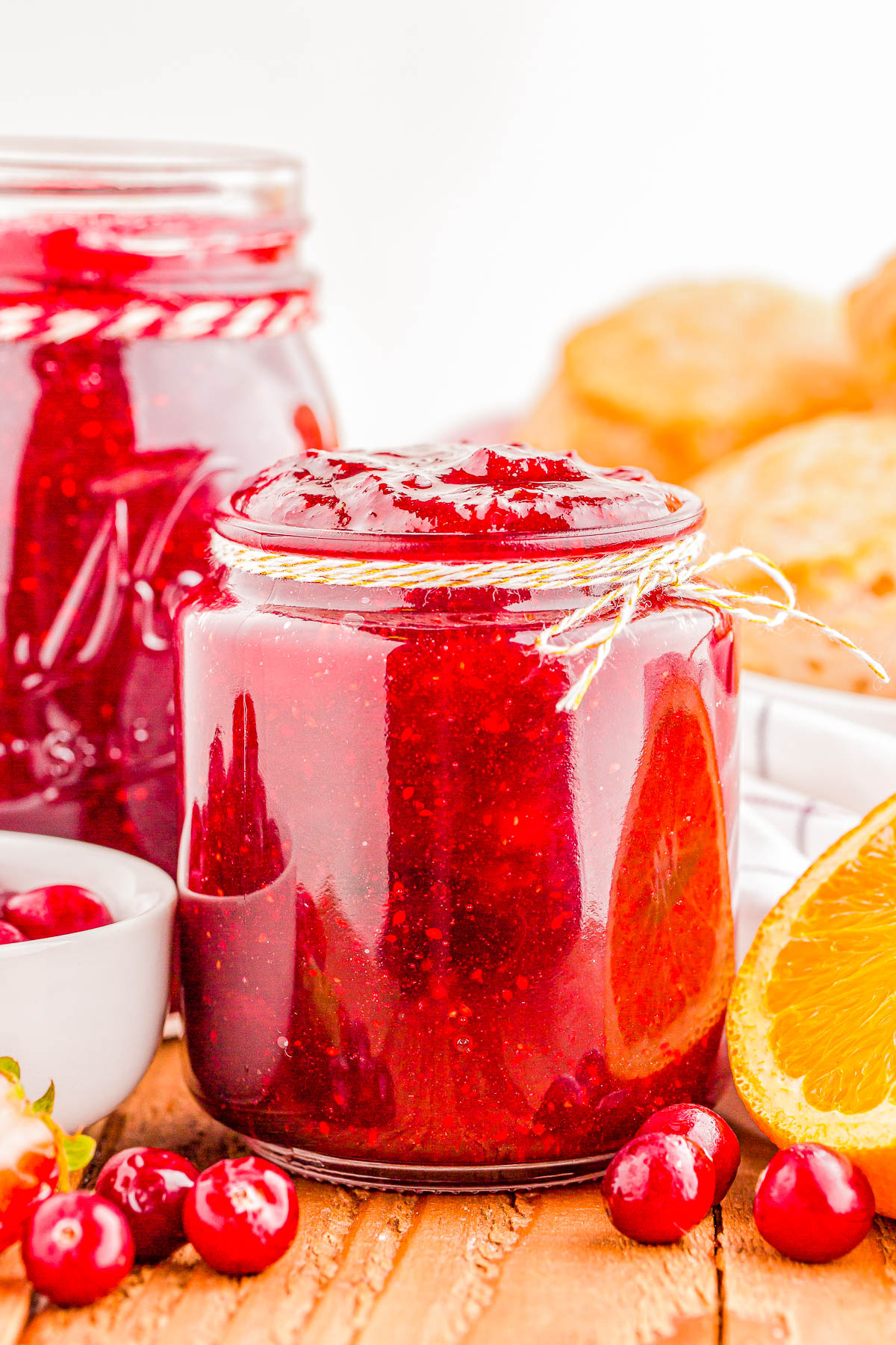 A jar of cranberry sauce surrounded by fresh cranberries, orange slices, and pastries on a wooden surface.