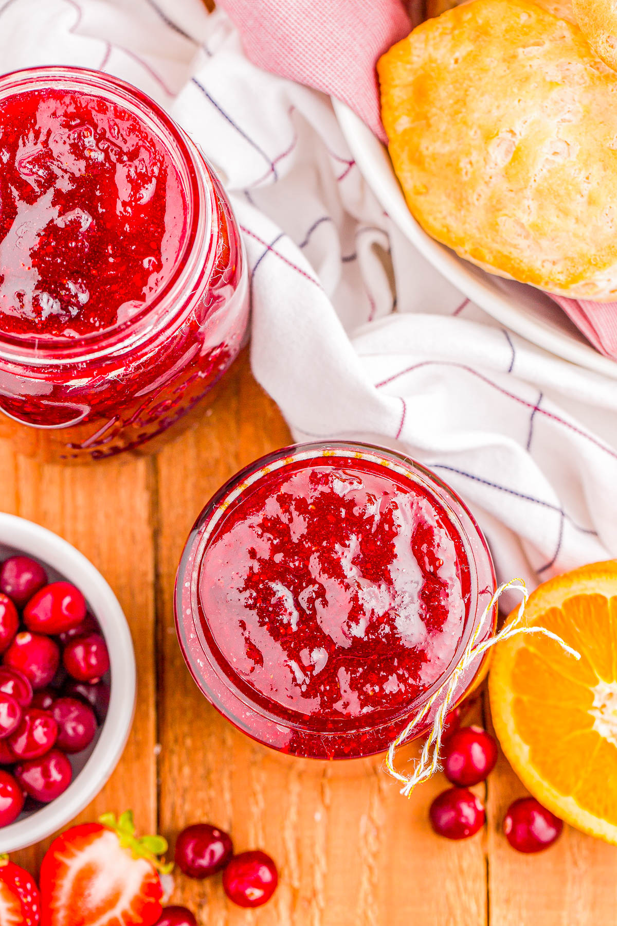 Jars of cranberry sauce, surrounded by cranberries, an orange slice, a biscuit, and a striped cloth, on a wooden surface.