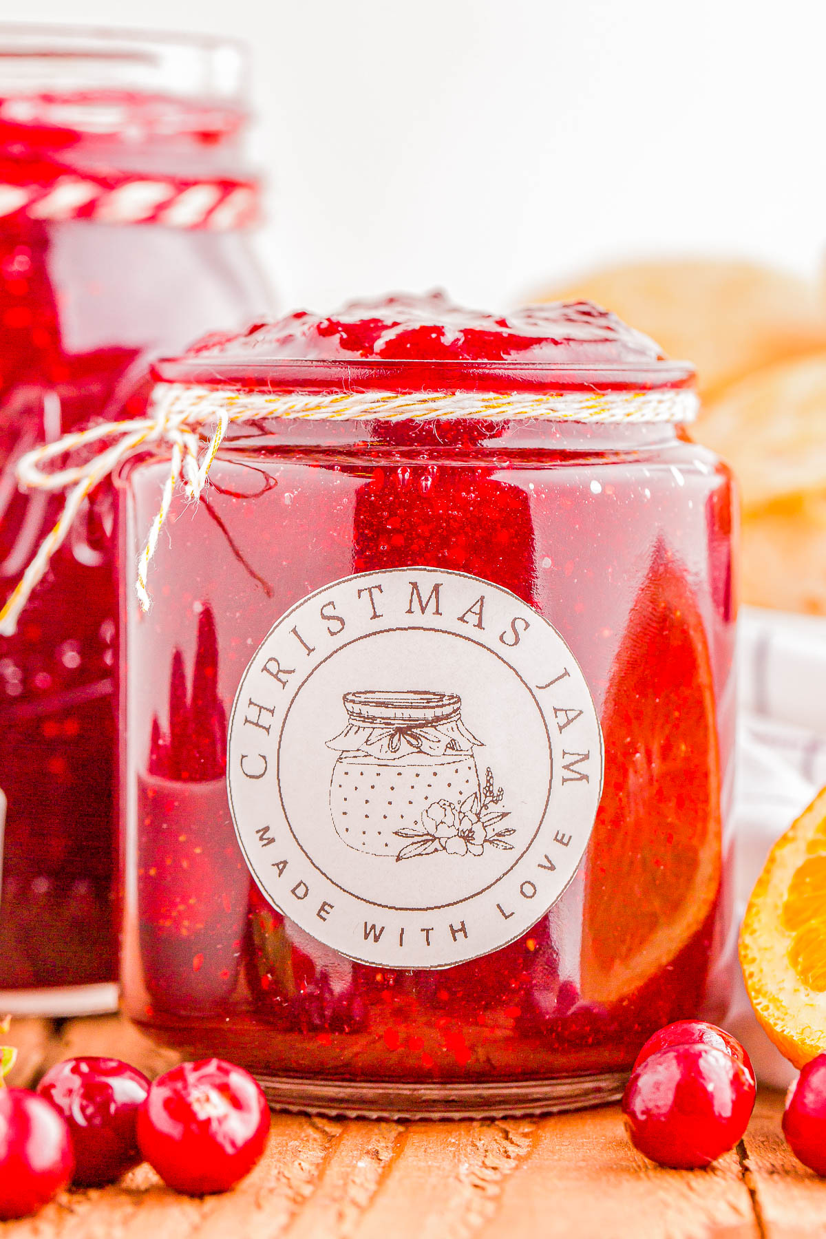 A jar of red Christmas jam labeled "Made with Love," surrounded by cranberries and orange slices.