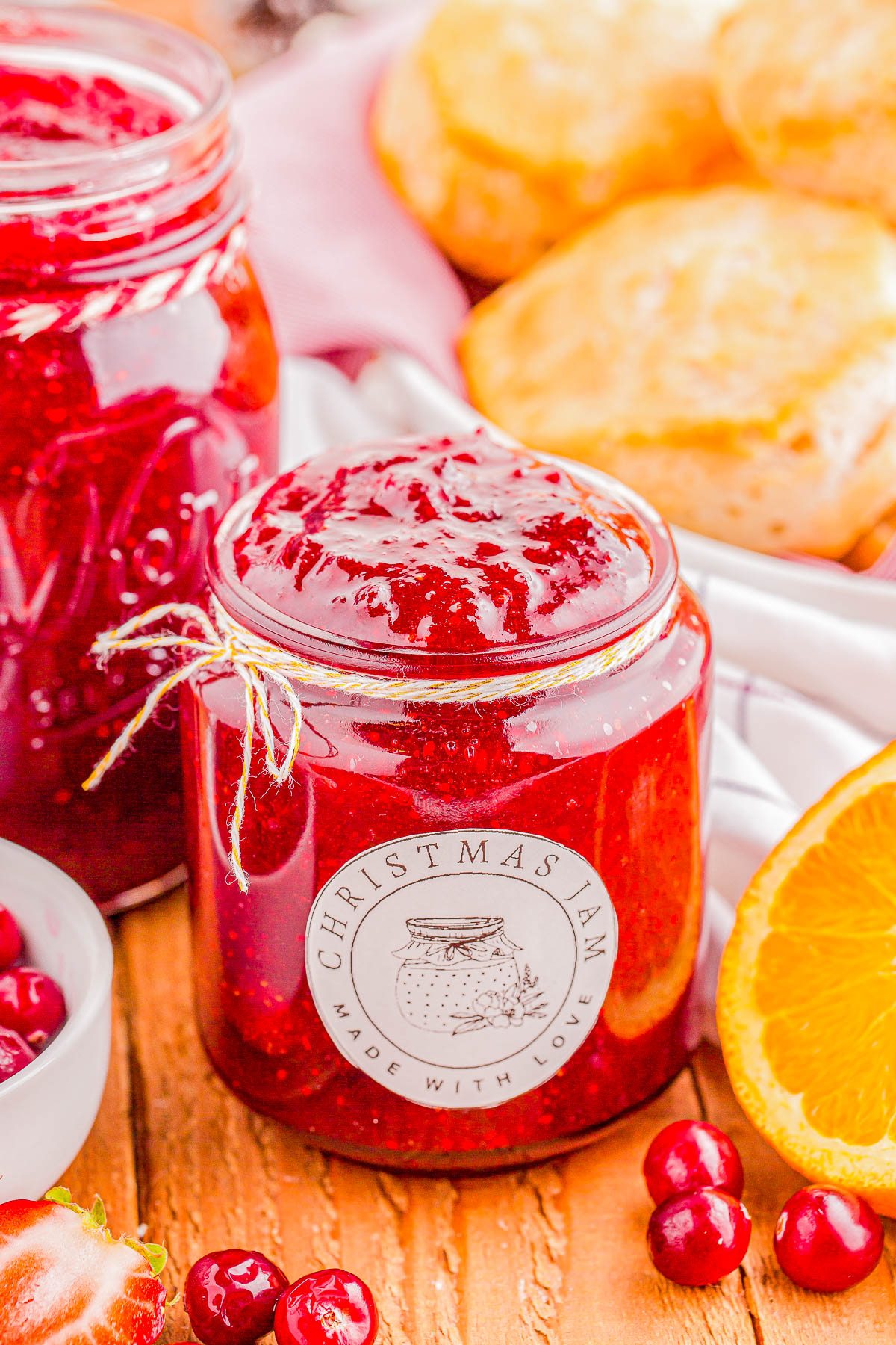 Jar of red Christmas jam on a wooden surface, surrounded by cranberries, orange slices, and scones.