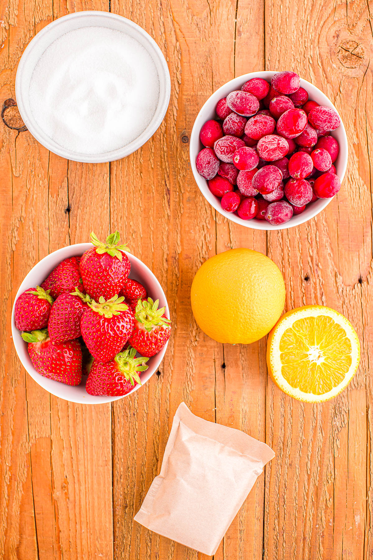 Strawberries and cranberries in bowls, a whole and halved orange, a bowl of sugar, and a small brown packet on a wooden surface.