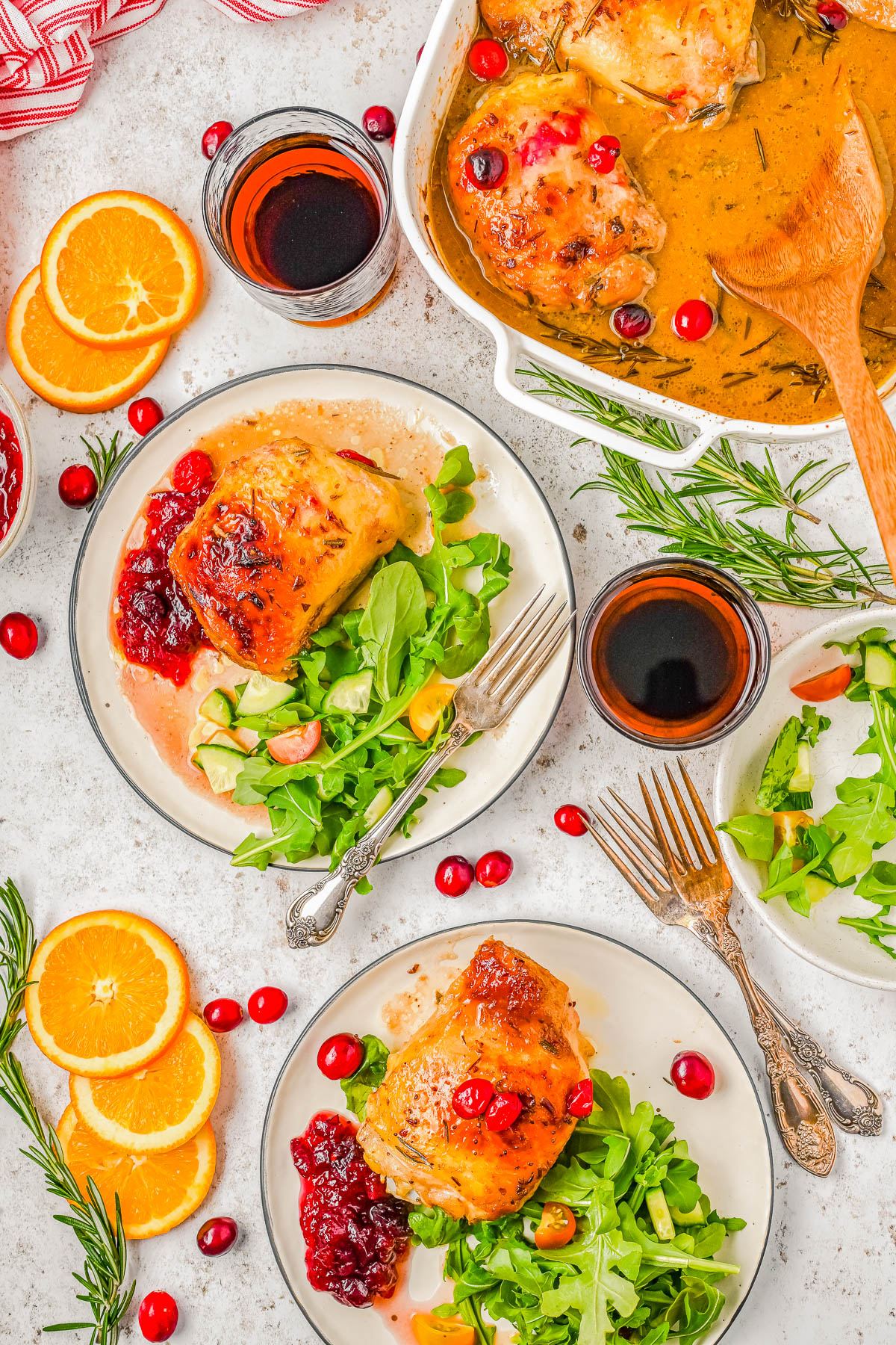 Plate of roasted chicken with cranberry sauce and arugula salad, surrounded by orange slices, cranberries, rosemary, and a baking dish with more chicken. Glasses of juice and utensils nearby.