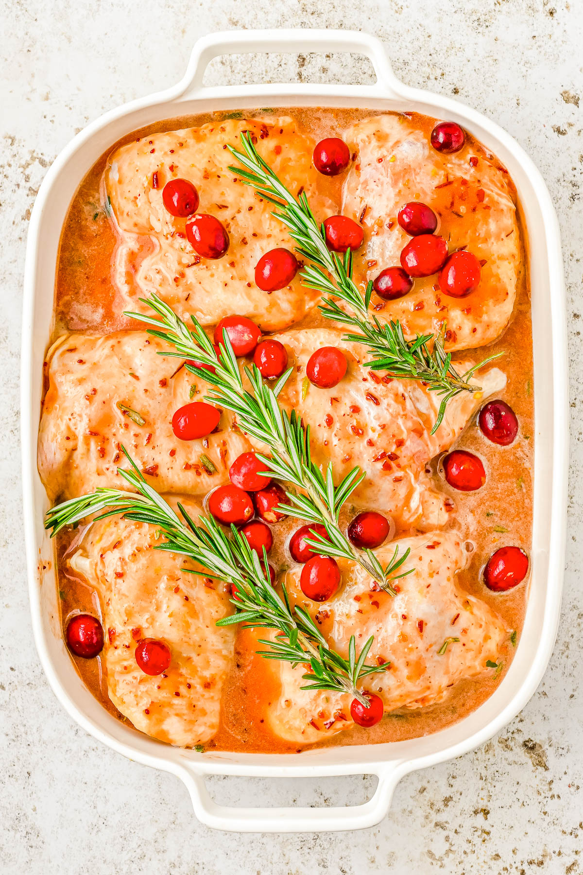 A baking dish with seasoned chicken pieces, topped with fresh rosemary and cranberries, in a marinade.