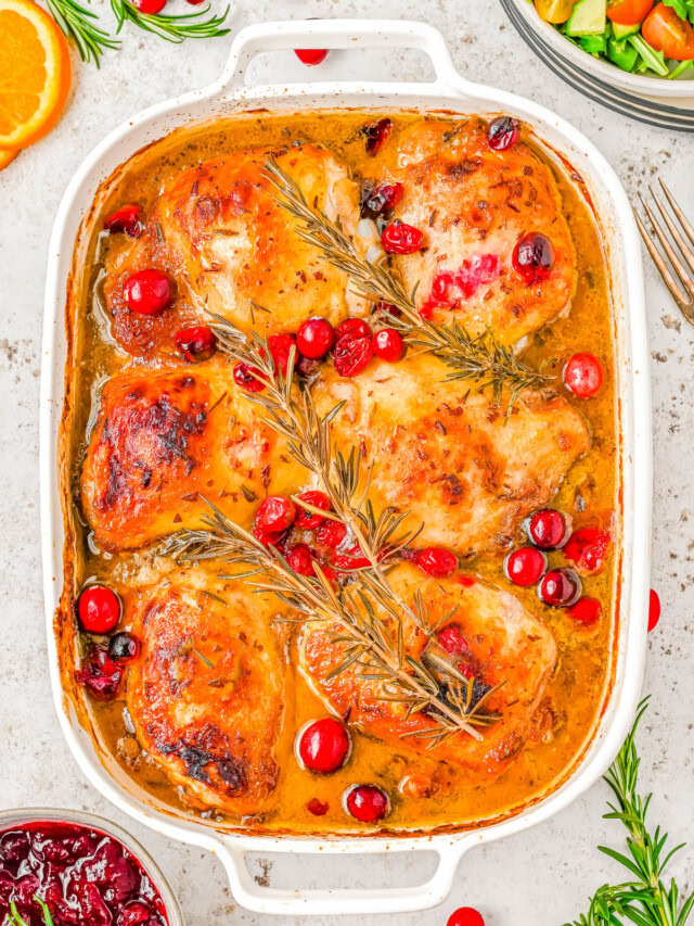 Baked chicken thighs in a casserole dish with cranberries and rosemary, garnished with orange slices.