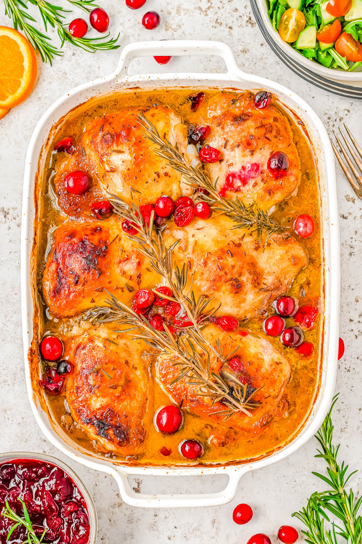 Baked chicken thighs in a casserole dish with cranberries and rosemary, garnished with orange slices.