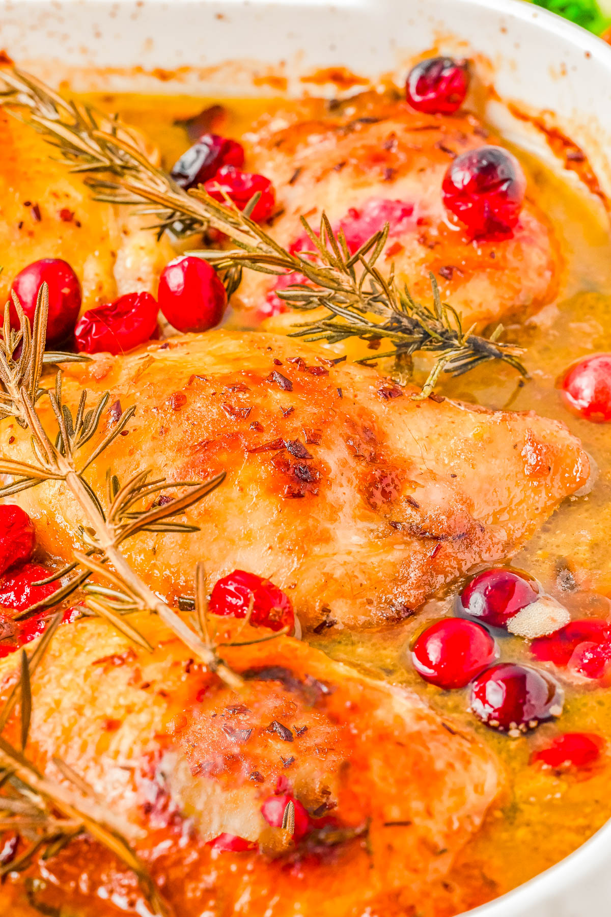 Baked chicken with rosemary, cranberries, and a golden glaze in a white dish.