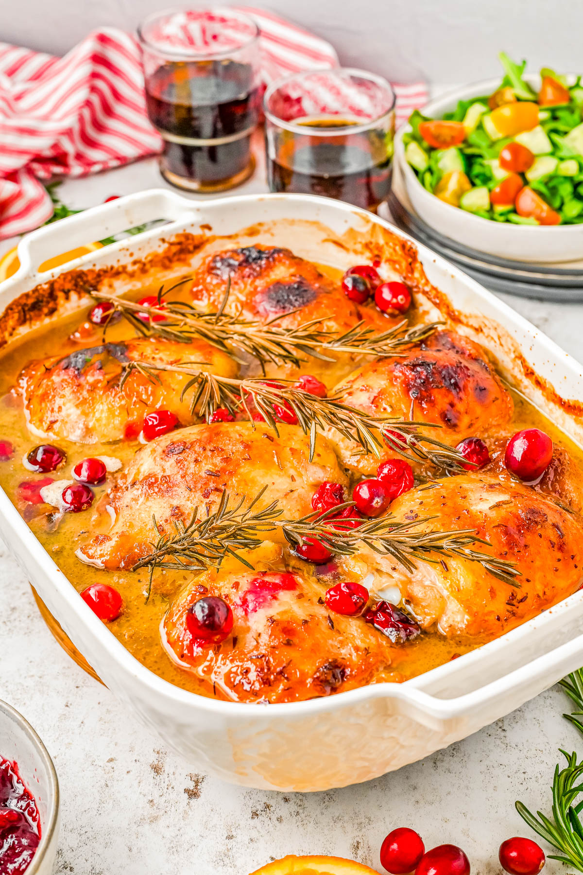 Baked chicken thighs topped with cranberries and rosemary in a casserole dish. Two glasses of dark beverage and a bowl of salad with cucumbers and lettuce are in the background.