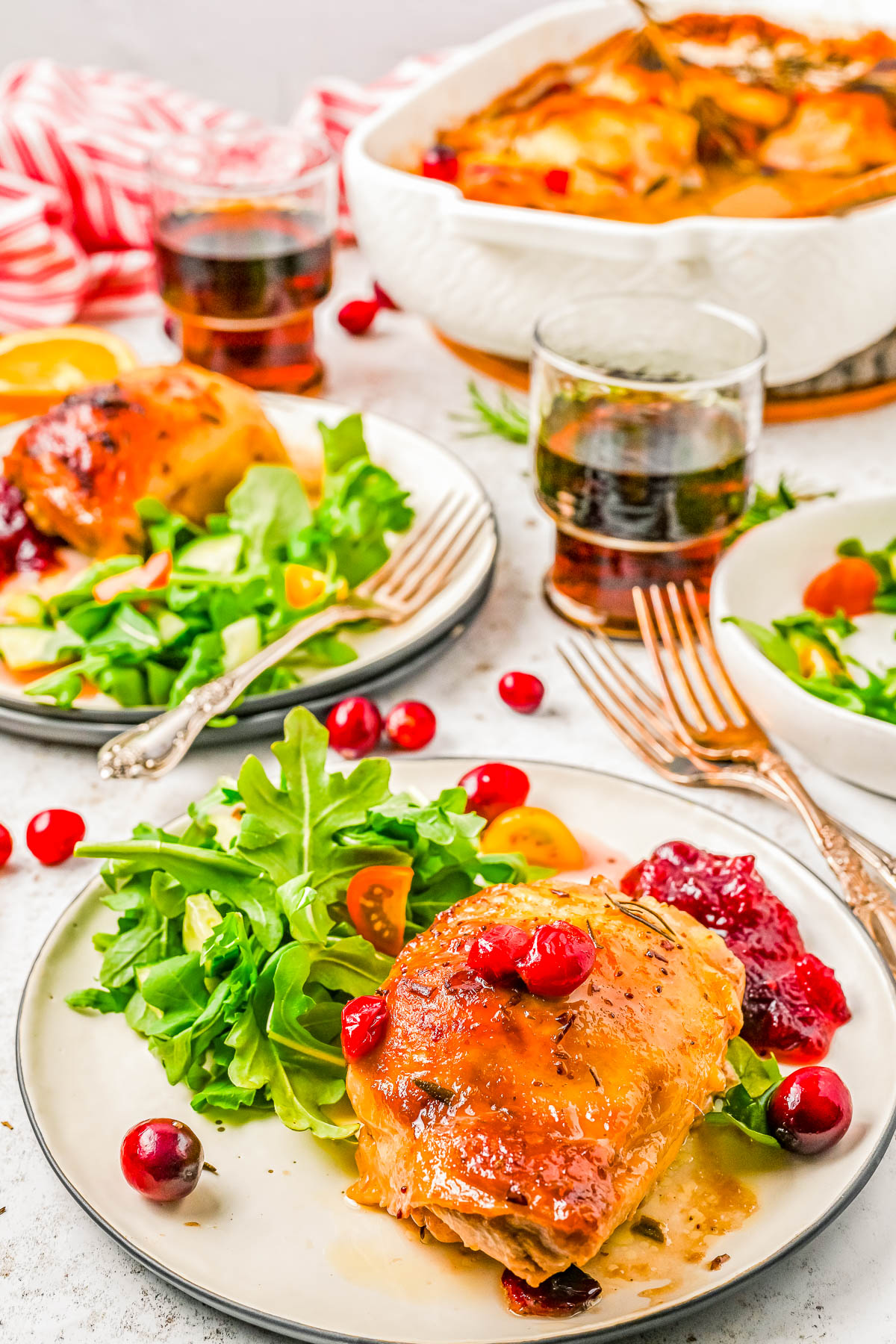 Plates of roasted chicken garnished with cranberries, served with fresh green salad and glasses of dark beverage on a white table.