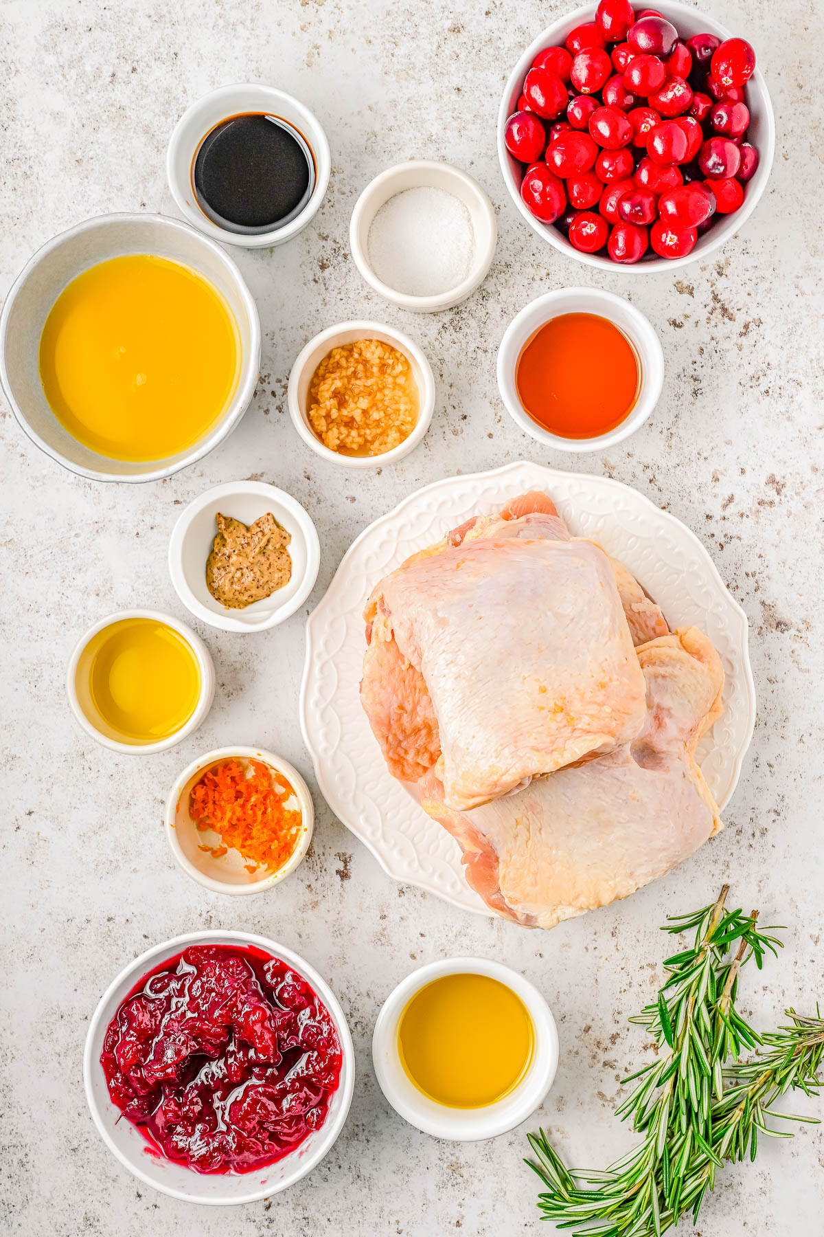 Raw chicken thighs on a plate surrounded by small bowls of various ingredients, including cranberries, rosemary, sauces, and seasonings on a light background.