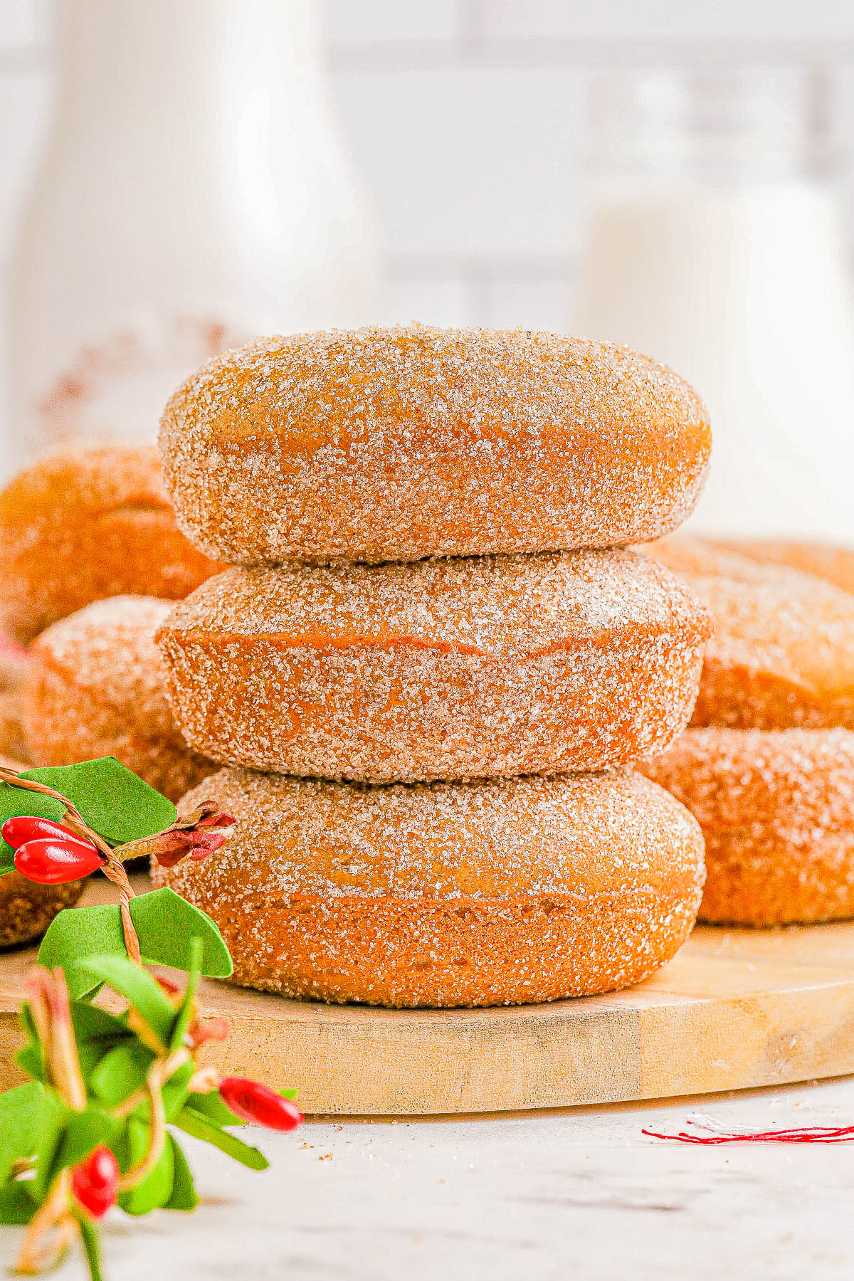 Three sugar-coated doughnuts stacked on a wooden board with more doughnuts and a milk bottle in the background, alongside a plant with small red flowers.