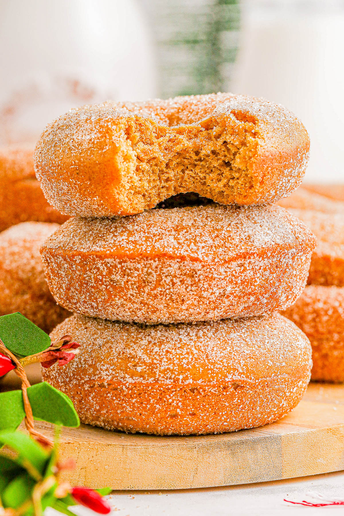 Three sugar-coated donuts stacked; the top one has a bite taken out. A small piece of greenery and red flowers are at the bottom left.