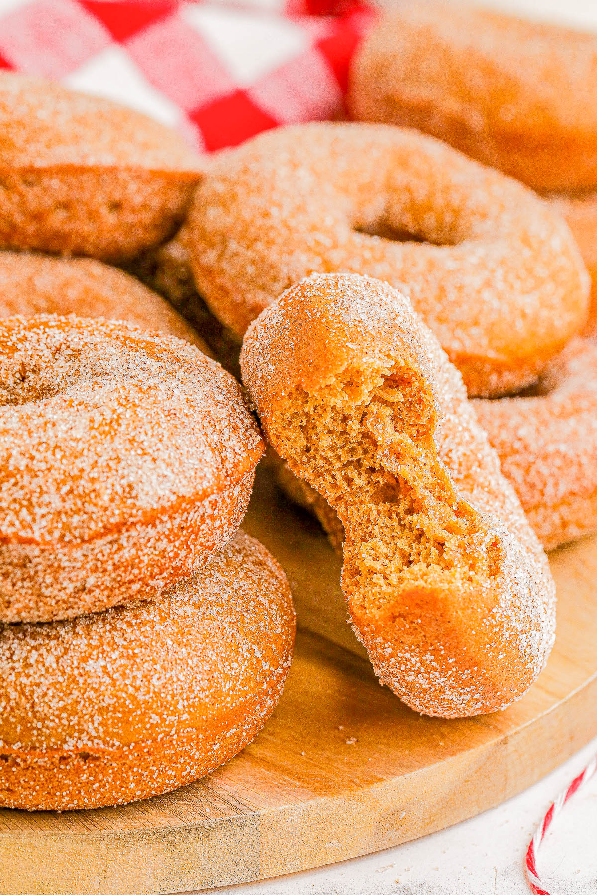 A stack of sugar-coated donuts on a wooden board, with one donut having a bite taken out of it. Red and white checkered cloth in the background.