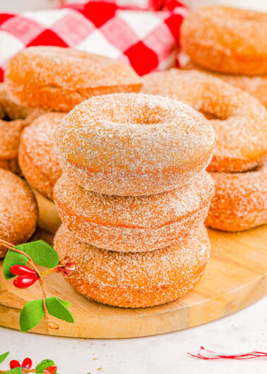 A stack of three sugar-coated donuts on a wooden board, surrounded by more donuts and sprigs with red berries.