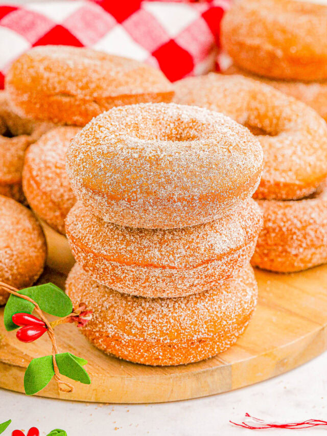 A stack of three sugar-coated donuts on a wooden board, surrounded by more donuts and sprigs with red berries.