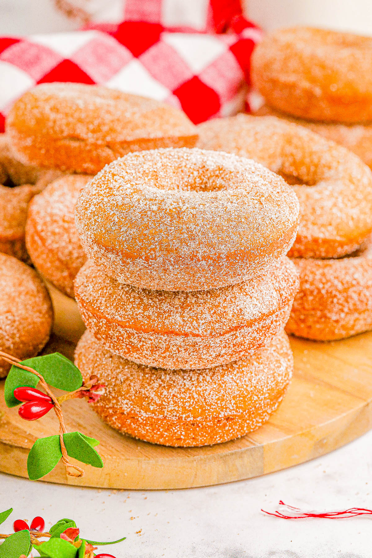 A stack of three sugar-coated donuts on a wooden board, surrounded by more donuts and sprigs with red berries.