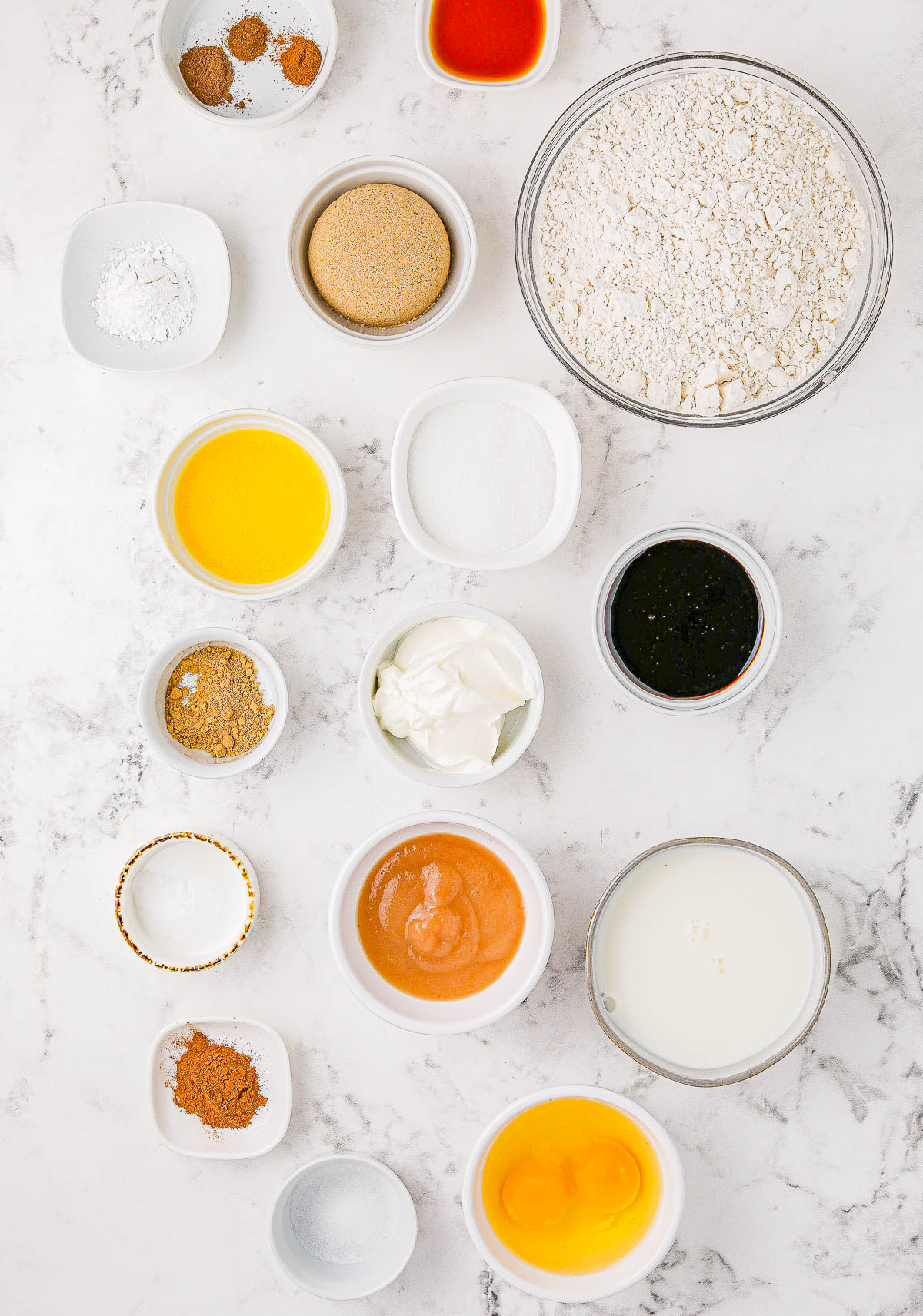 Assorted baking ingredients including flour, sugar, eggs, spices, milk, molasses, and butter arranged on a marble surface.