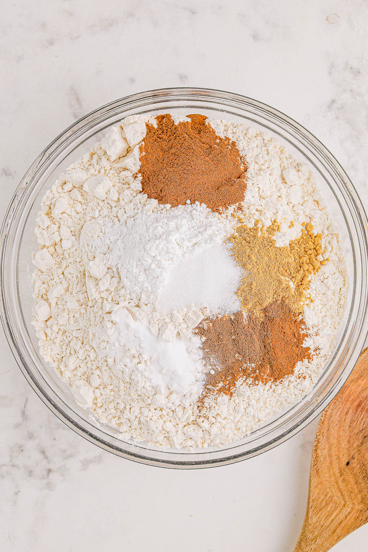 A bowl containing flour, cinnamon, salt, baking powder, and other spices on a marble surface with a wooden spoon beside it.