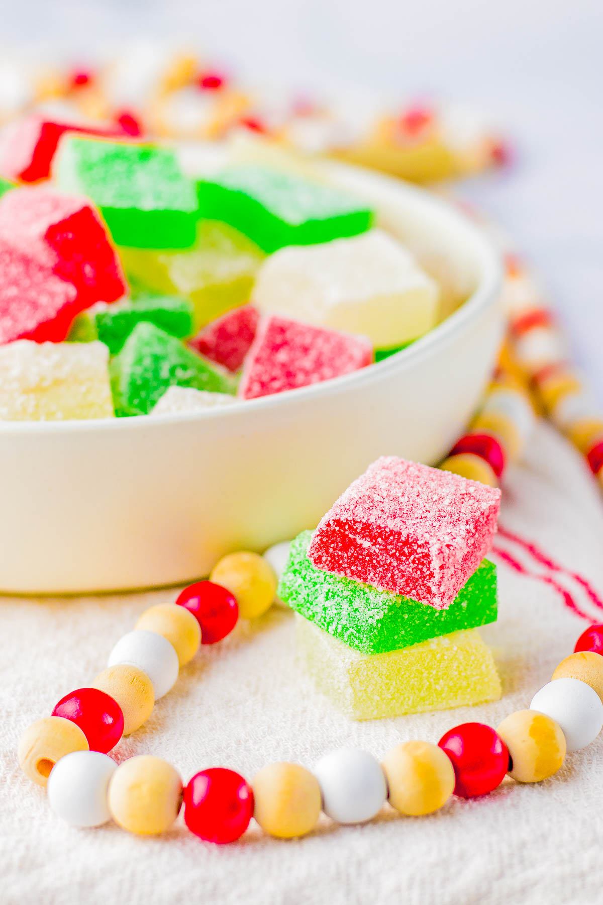 A bowl of red, green, and yellow sugar-coated jelly candies with a wooden bead necklace nearby on a white surface.